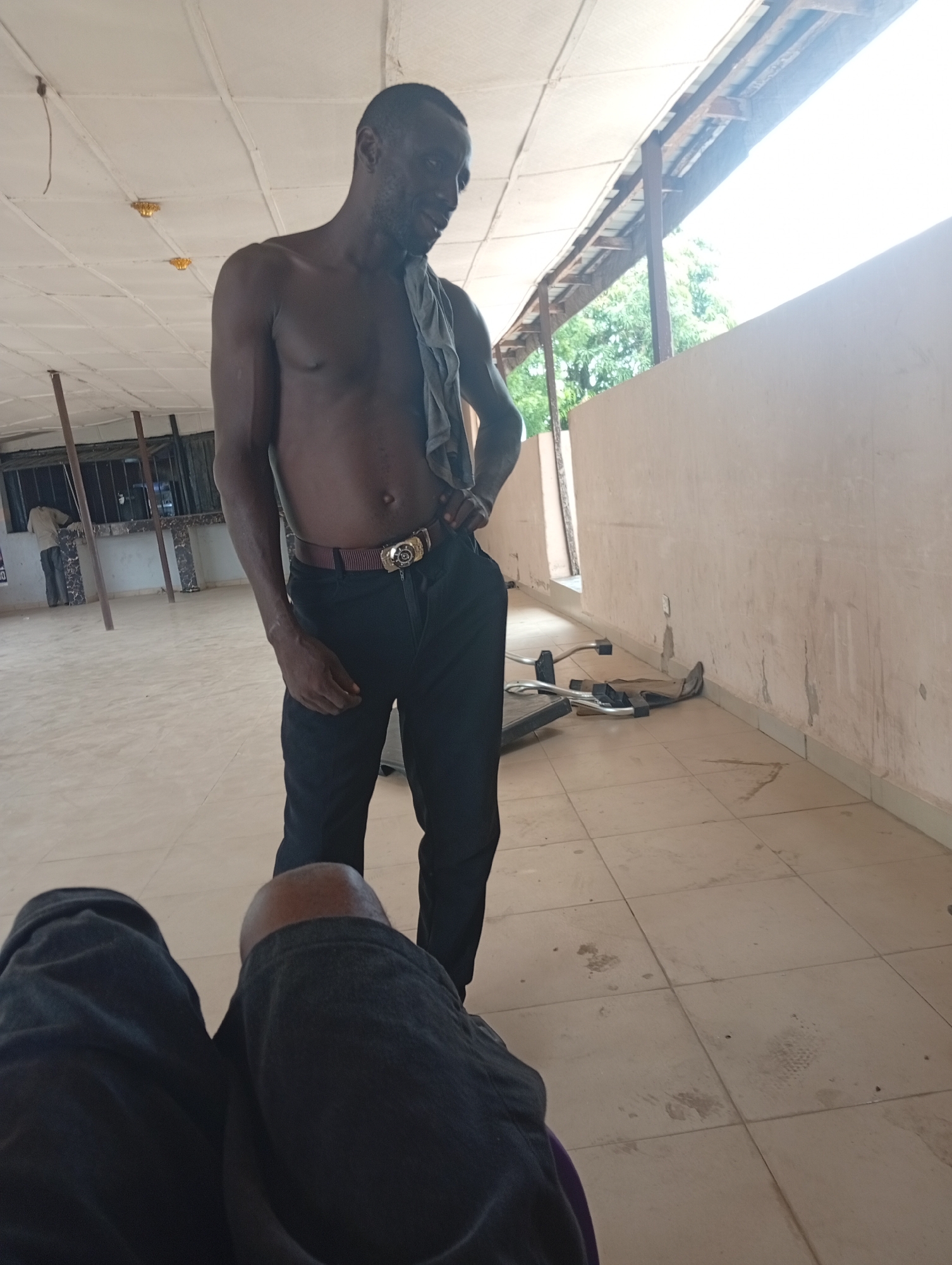 A man stands shirtless in an indoor space, wearing black pants with a gold-buckled belt. A gray cloth drapes over his shoulder. His right hand rests on his hip. Another person's legs, clad in dark fabric, are visible in the foreground, suggesting a seated perspective. The room has a tiled floor and off-white walls. Outside, seen through an opening, are trees and a structure with a corrugated roof. Another man is visible in the background, standing at what appears to be a window. The city is Adankolo, Nigeria.