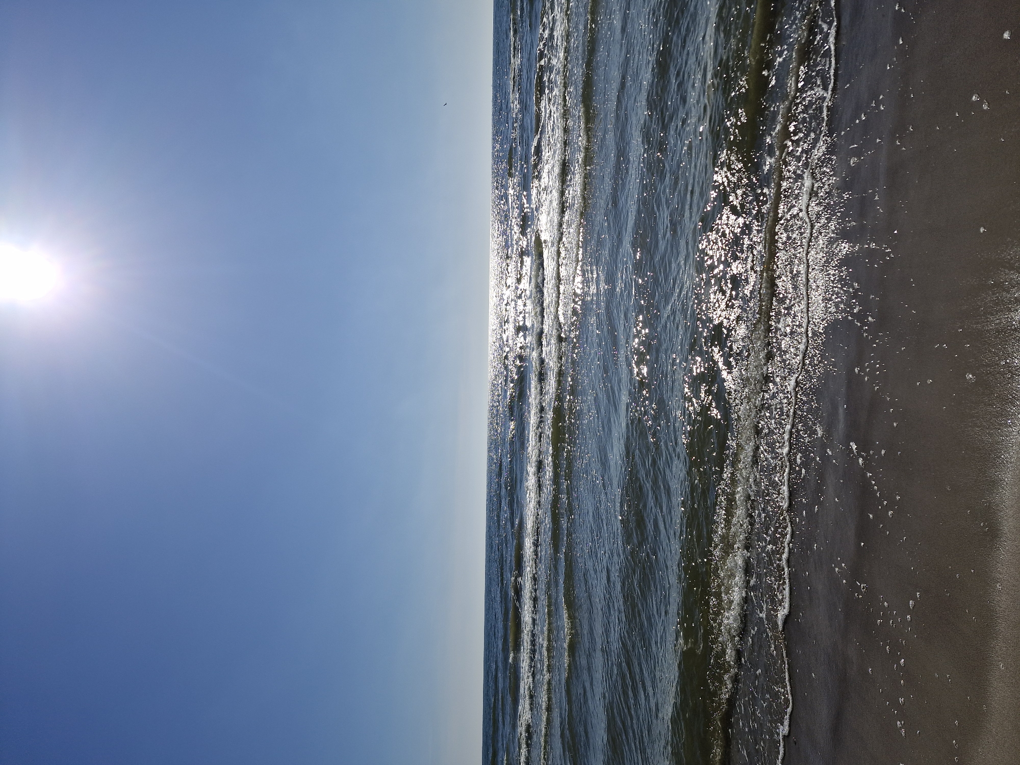 The image depicts an outdoor coastal scene under a clear blue sky, with the sun positioned in the upper right quadrant, casting intense light and reflections across the water. The upper half of the frame is dominated by a cloudless, light blue sky. A bright, circular sunburst is visible in the upper right, generating prominent lens flare radiating downwards and to the left. The middle section of the image features the ocean, characterized by dark blue-green water with numerous waves extending horizontally. The direct sunlight creates a shimmering, speckled pattern of bright reflections across the water's surface, particularly in the mid-distance towards the horizon. A minute, dark avian silhouette is discernible flying over the water in the far right section. The lower portion of the image displays a wet, dark brown sandy beach, with white foam and scattered bubbles marking the edges of the receding waves. The horizon line, appearing level, separates the sea from the sky approximately one-third of the way from the bottom of the frame. No human figures or specific man-made structures are present.