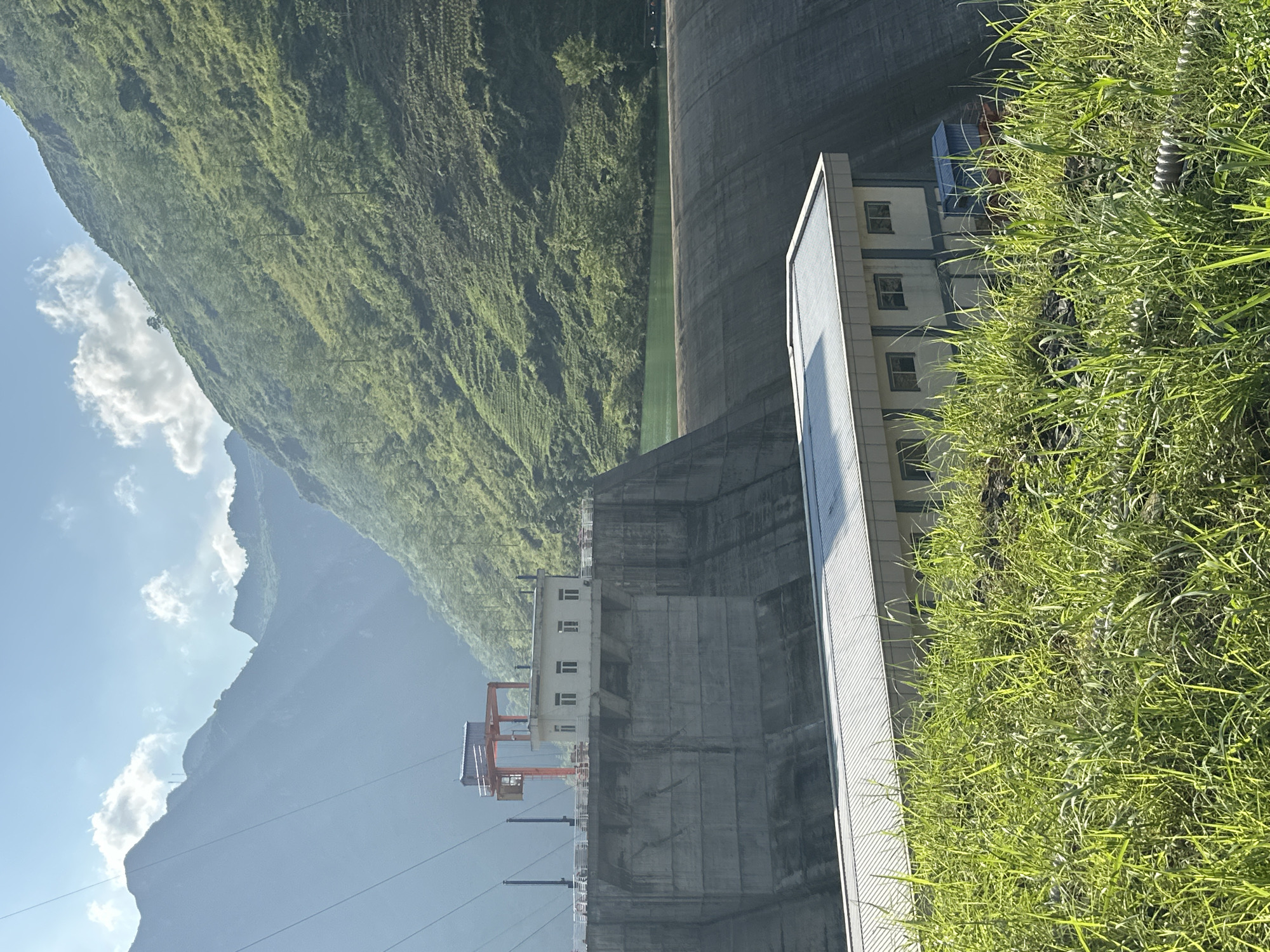 The image shows a hydroelectric dam in Sơn Vĩ Commune, Vietnam. The dam is constructed of concrete and spans a valley between green, mountainous terrain. On the right side of the dam, a rectangular building with multiple windows rises several stories. Thick green grass grows in the foreground. Power lines stretch across the lower part of the image. The sky is blue with scattered clouds.