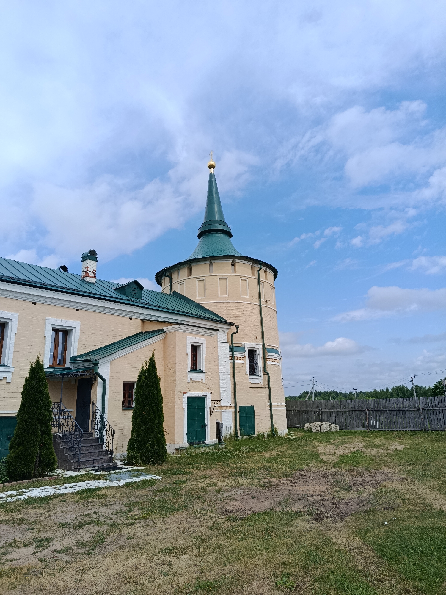 The image depicts the Nikolo-Radovitskiy Monastery in Radovitsy, Russia. The monastery features a prominent, pale yellow, octagonal tower with a green conical roof topped with a golden cross. Attached to the tower is a long, low building with a green roof, also painted pale yellow. The building has several windows with dark wooden frames and green doors. Two tall, dark green coniferous trees stand in front of the building, flanking a set of stone stairs leading to an entrance. The ground in the foreground is covered with dry, yellowed grass and patches of bare earth. In the background, a weathered wooden fence runs horizontally, with a line of green trees beyond it. The sky above is a bright blue, with scattered white clouds.