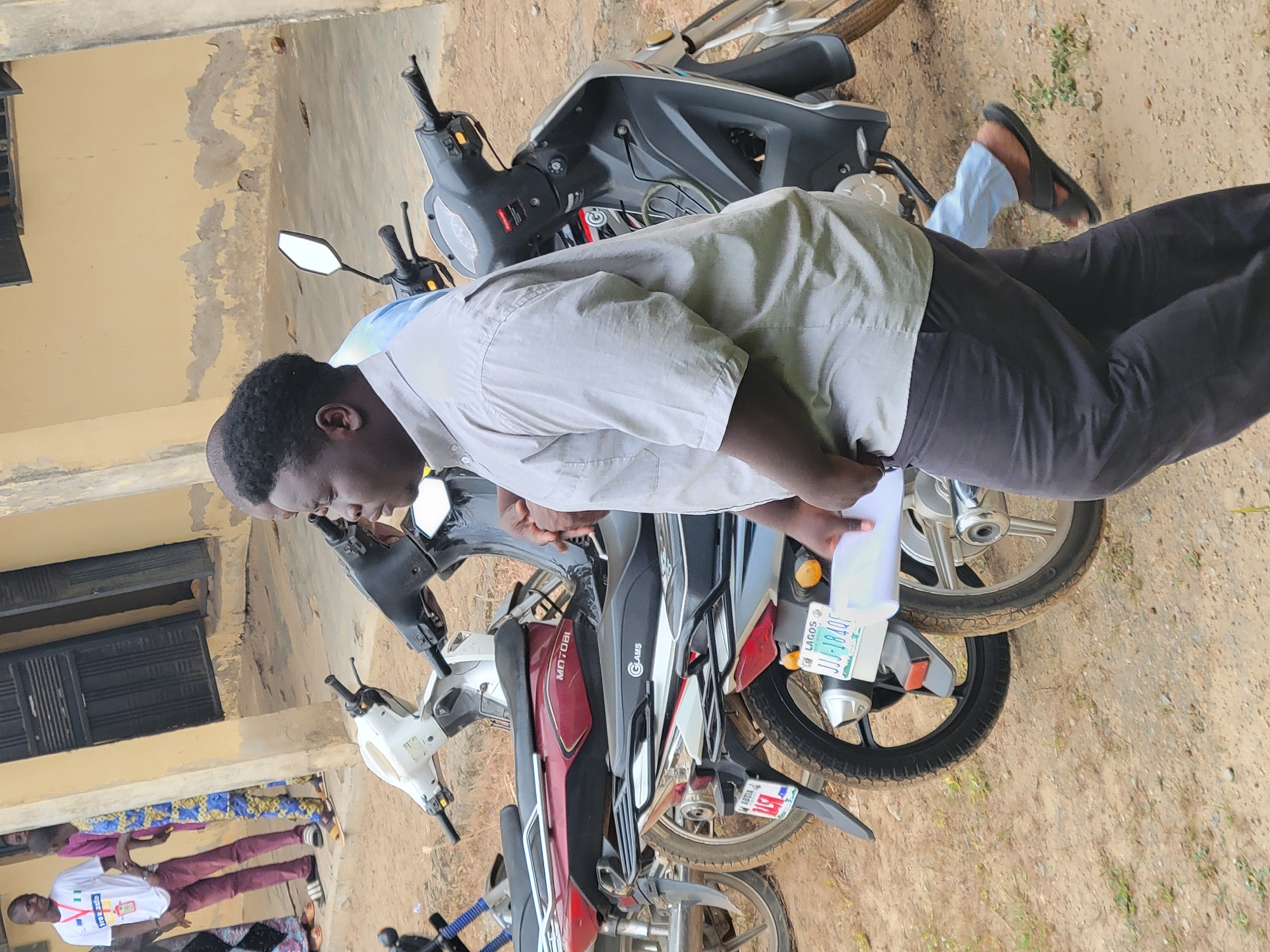 The image captures an outdoor scene, likely in Keffi, Nigeria, depicting a man focused on paperwork among several parked motorcycles. The main subject is a dark-skinned man, standing with his head bowed, intently examining white documents he holds in his hands. He wears a light grey short-sleeved collared shirt over a lighter blue layer and dark trousers.
He is positioned in front of a line of motorcycles and scooters. Visible vehicles include a grey and black scooter, a white scooter, a dark red motorcycle with "MOTOBI" emblazoned on its seat, and another motorcycle displaying "GLAMS" on its side. A distinctive green and white license plate on one of the motorcycles clearly reads "LAGOS JJJ-18AE".
In the background to the left, several other people are partially visible, suggesting a small gathering or queue. One individual wears a white t-shirt with fragmented text like "OSSE..." and "NCE" or "MCE" visible, paired with burgundy trousers. Behind the people and vehicles is a light yellow or tan building with noticeable patches of peeling paint, indicating wear. A dark, possibly wooden, door or shutter is visible on the building's facade. The ground is unpaved, appearing dry, dusty, and sandy, with scattered small green plants. The scene takes place during daylight hours, under natural light, and conveys a focused activity, possibly related to vehicle registration or administration.