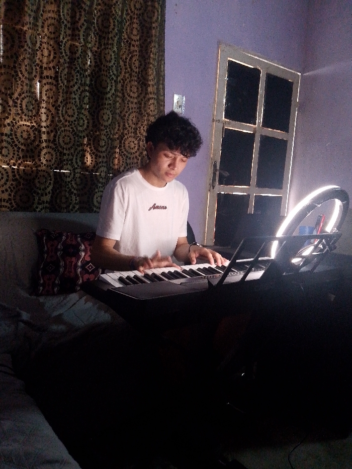 A young man with curly hair sits at a keyboard, wearing a white t-shirt with text. Behind him is a patterned curtain and a white door with dark rectangular panes. To the right, a ring light shines brightly. The keyboard and its stand are partially obscured by shadows. A decorative pillow rests on a couch behind the man. The room appears to be dimly lit, with a purple-ish hue to the walls.