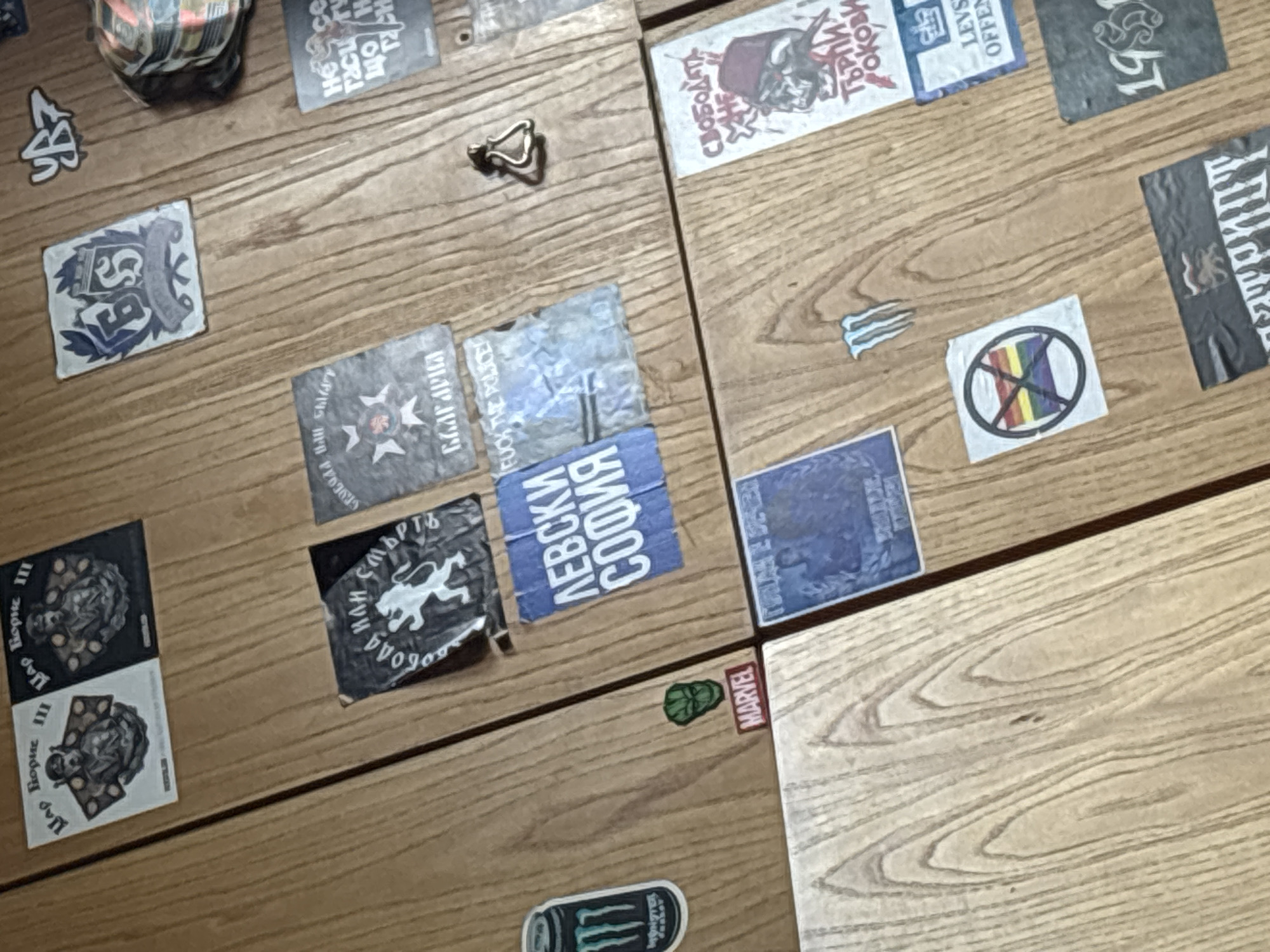 The image displays a light-colored wooden surface, possibly a desk or a large panel, covered with a diverse collection of stickers. The surface has visible seams, dividing it into sections.
The stickers reflect various interests and affiliations. Many feature Cyrillic text, strongly suggesting a Bulgarian context. These include two identical stickers of "ЦАР БОРИС III" (Tsar Boris III) with a portrait, a sticker with "БЪЛГАРИЯ" (Bulgaria) and an emblem, and a blue sticker for "ЛЕВСКИ СОФИЯ" (Levski Sofia), a Bulgarian football club. Another prominent sticker displays "СВОБОДА ИЛИ СМЪРТЬ" (Freedom or Death) with a lion, a well-known Bulgarian revolutionary motto. A partially visible sticker reads "СВОБОДАТА НЕ ТЪРПИХ..." (Freedom does not tolerate...). Additional text includes "LEVS OFFEN" and "1955".
Other stickers include: a "FUCK THE POLICE" text, a rainbow flag with a diagonal line through it (indicating opposition to LGBTQ+ rights), a small green Hulk head, and a "MARVEL" logo. A partial Monster Energy drink logo is also visible. Near the top left, a crumpled can is partially shown. A small, metallic, abstract object, possibly a piece of jewelry or a decorative item, rests on the wood. The overall scene is a static display of personal expression through stickers on a wooden surface in an indoor setting. The time of day and weather are not discernible.