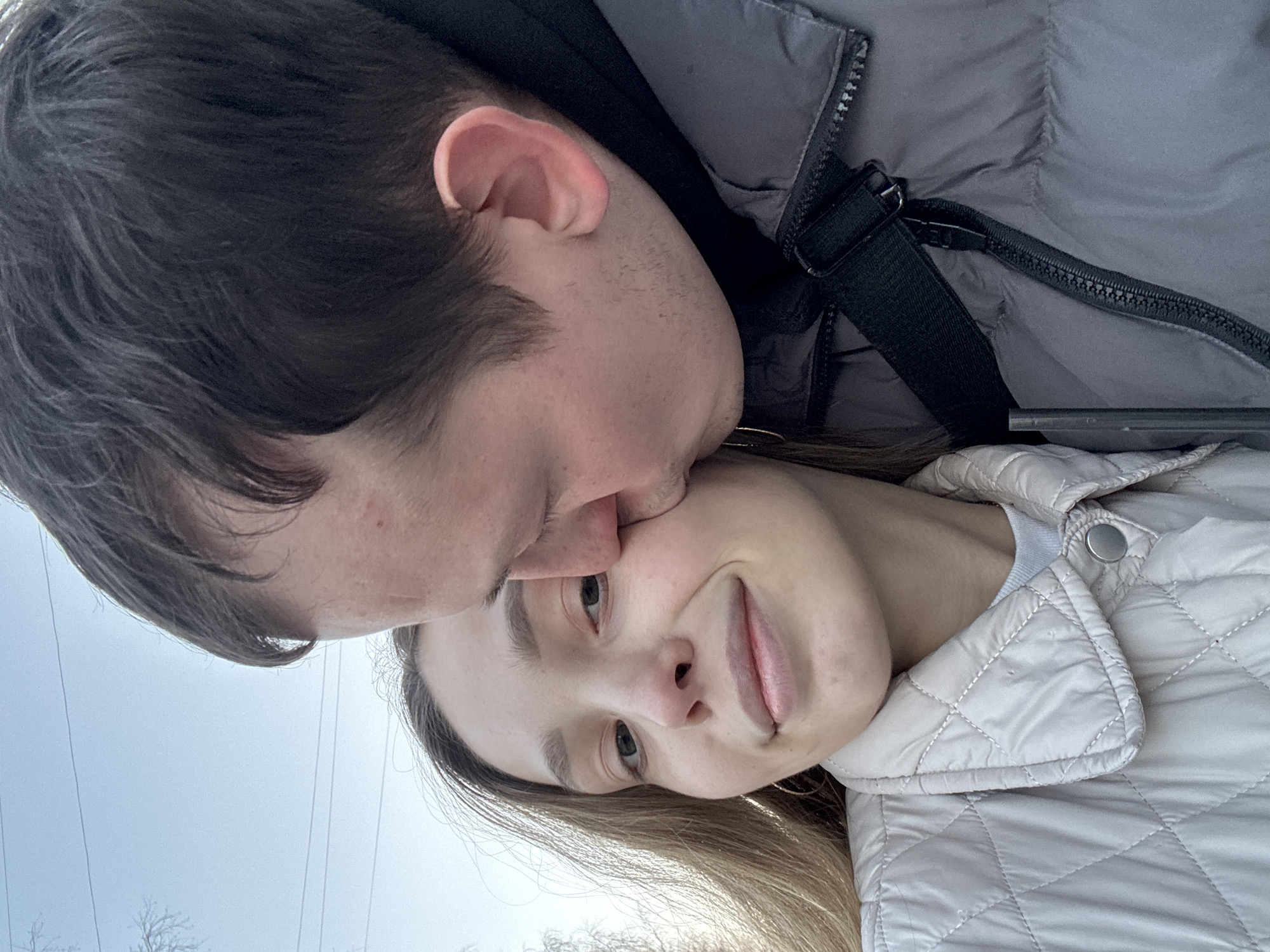 The image displays two individuals, a female and a male, outdoors in Chaikovsky, Russia. The female, with long light-brown hair, occupies the left half of the frame, wearing a light beige quilted jacket and looking towards the viewer with a slight smile. A thin gold hoop earring is visible on her right ear. The male, positioned to her right, has short dark hair and is wearing a dark grey puffer jacket with a black strap over his shoulder. He is actively kissing the female on her right cheek, with his lips pressed against her skin. The background consists of a clear, light blue sky, with sparse bare tree branches and multiple utility wires visible in the upper left quadrant.
