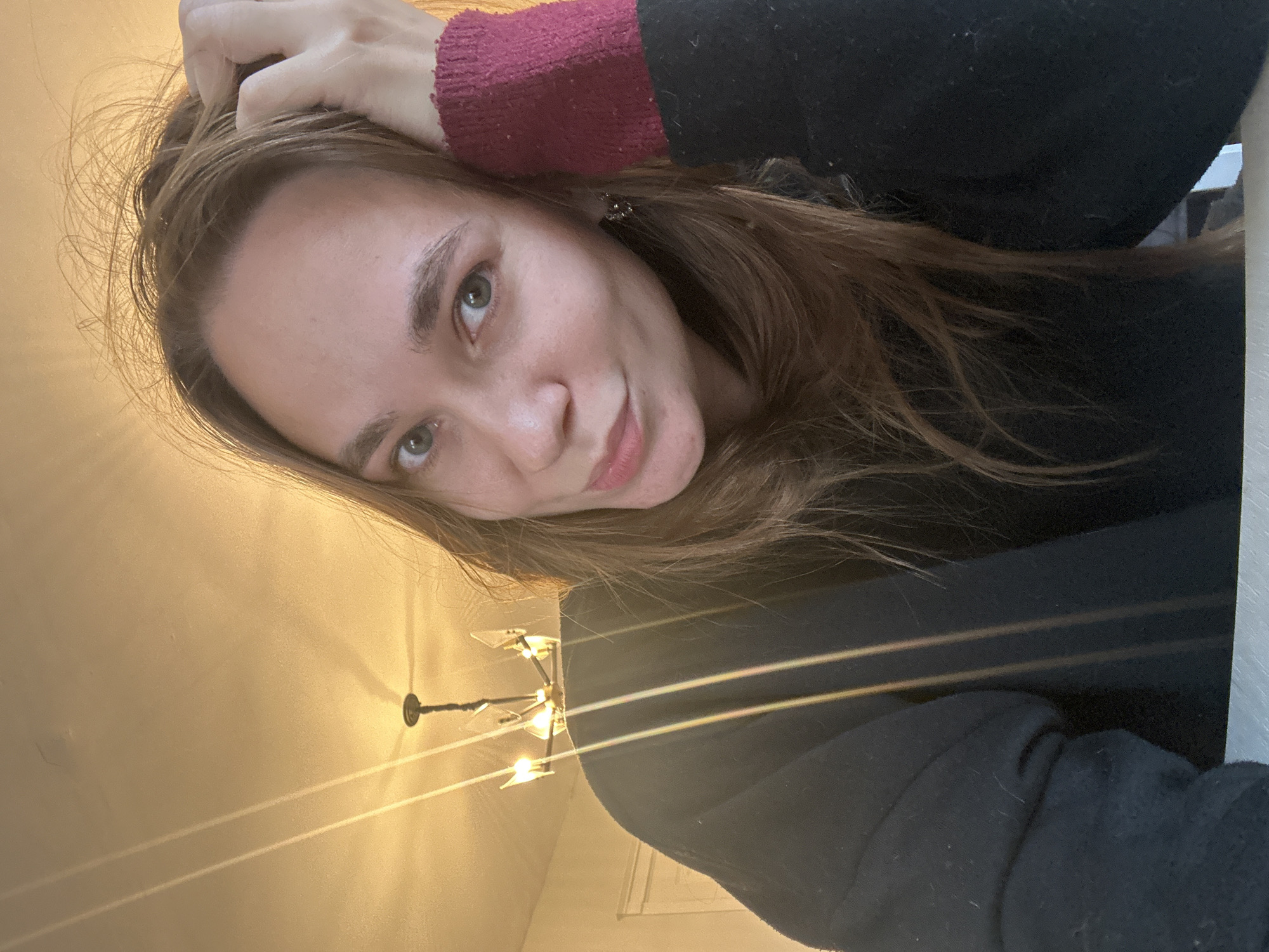 An indoor, head-and-shoulders portrait of a female subject, located in the city of Kharnavy, Russia. The subject has light brown, slightly disheveled hair and greenish-hazel eyes, looking directly at the camera with a subtle smile. She wears a black long-sleeved garment. Her left arm, featuring a maroon-colored knitted cuff, is raised, with her left hand positioned within her hair on the right side of the frame. In the upper background, a multi-bulb ceiling chandelier with geometric glass shades casts defined shadows on the warm-toned ceiling. Vertical lens flare lines are present on the left and center of the image. A faint light-colored architectural detail, possibly a doorframe, is visible on the far left, and a white surface forms the bottom edge of the frame.