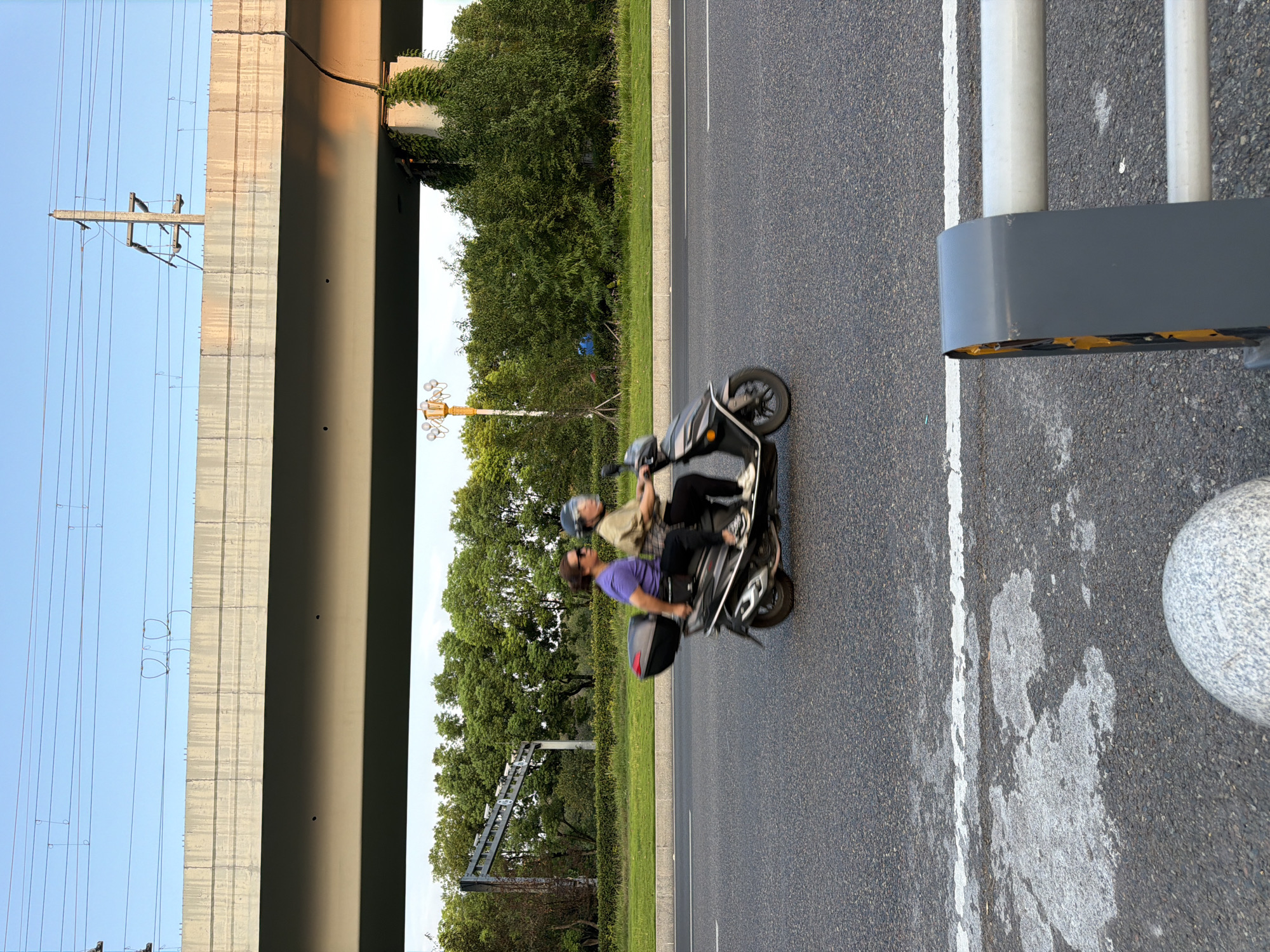 Two individuals are riding a scooter on an asphalt road in Jiangjin, China, during daylight. The scooter is moving from left to right in the frame. The person in the front is wearing a light-colored shirt and a helmet, and appears to be driving. The person in the back is wearing a purple shirt and sunglasses, and is looking forward. The scooter has a black storage box mounted behind the rear seat.
The road is paved with asphalt and has white lane markings. In the background, there is a large concrete overpass supported by pillars. Vegetation, including trees and bushes, lines the side of the road and the area under the overpass. A yellow lamppost with multiple light fixtures is visible among the trees. Overhead power lines and support structures are visible above the overpass, against a clear blue sky. The lighting suggests it is either early morning or late afternoon. In the foreground, a portion of a metal barrier with grey and yellow elements is visible, along with a textured spherical object partially in the frame at the bottom left. The scene captures a moment of everyday transportation in an urban or semi-urban environment.