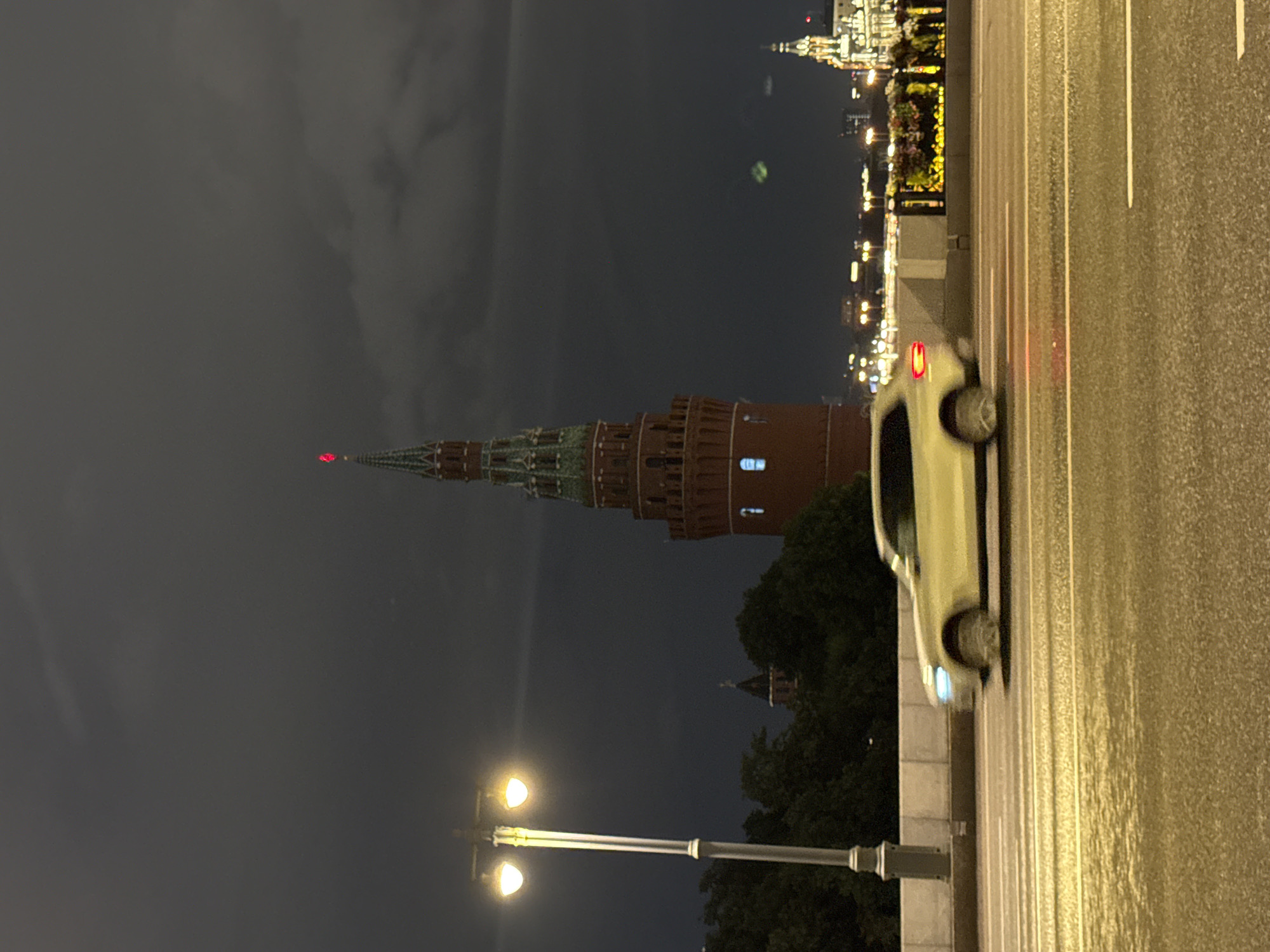 The image depicts a view of the Moscow Kremlin at night, with a car in motion on a bridge in the foreground. The Kremlin's Spasskaya Tower, a prominent landmark, is visible in the background, illuminated by a red star at its spire. The sky is dark and overcast, suggesting a cloudy or stormy night. A street lamp with two glowing bulbs stands to the left of the frame, casting light on the bridge's walkway. The bridge is wet, reflecting the lights of the city. The car, a white SUV, is blurred due to motion, indicating it is driving at speed. In the far background, the cityscape of Moscow is dotted with lights from buildings and bridges. The overall atmosphere is moody and urban, characteristic of a nighttime scene in a major city.