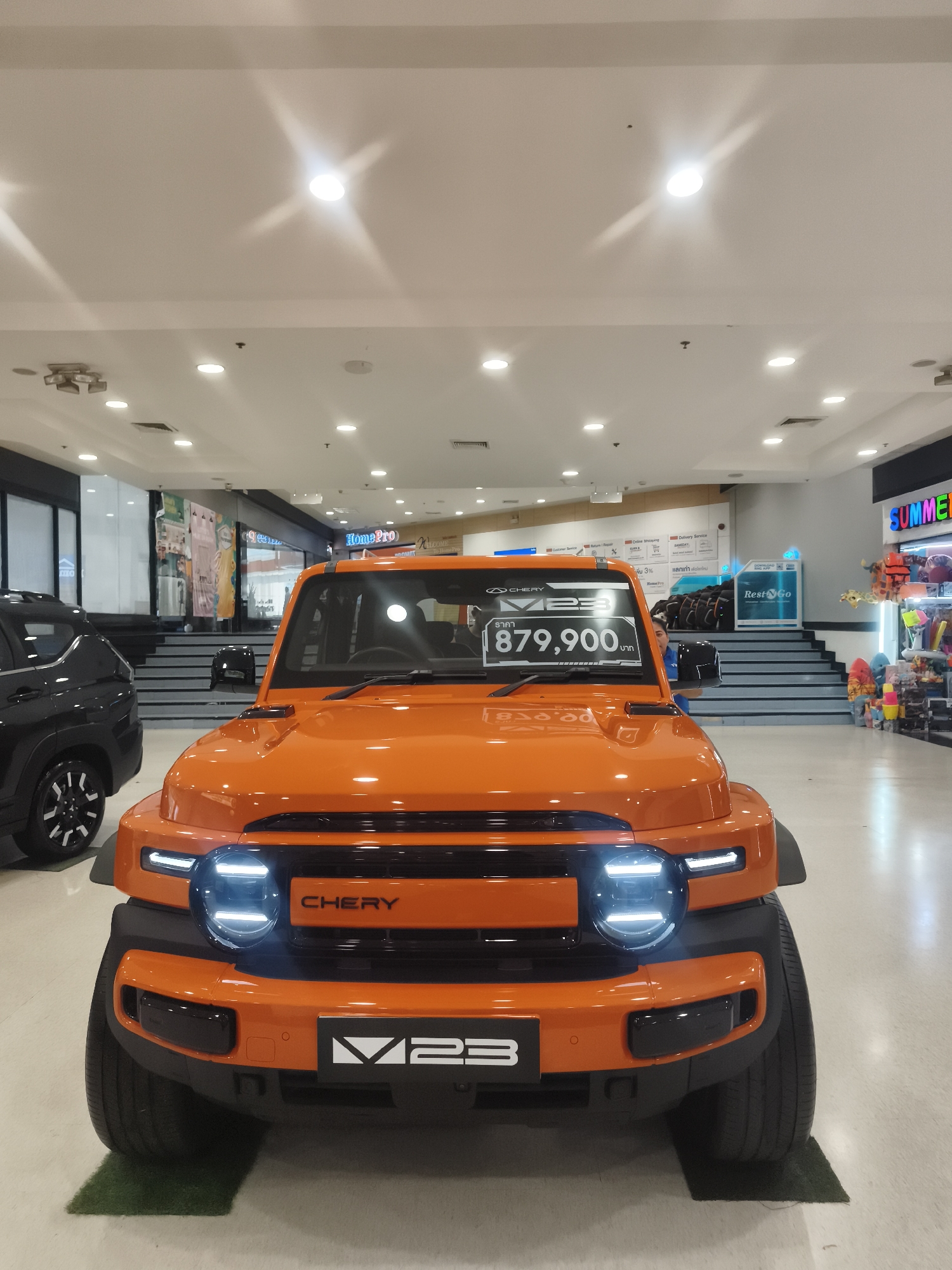 The image shows the front of an orange Chery M23 SUV on display in a brightly lit indoor shopping mall. The car has a prominent black grille with "CHERY" written across it, and round LED headlights. A price tag on the windshield reads "879,900 บาท". In the background, there are escalators leading up to other levels of the mall, with various storefronts visible, including "HomePro" and "SUMMER". To the left of the orange SUV, a black SUV is partially visible. The flooring appears to be polished concrete. The environment suggests a retail setting in a modern shopping complex. Given the visible signage and the architectural style, this scene is likely located in a shopping mall in a developed area of Ko Samui District, Thailand, possibly in Chaweng or Bophut.
