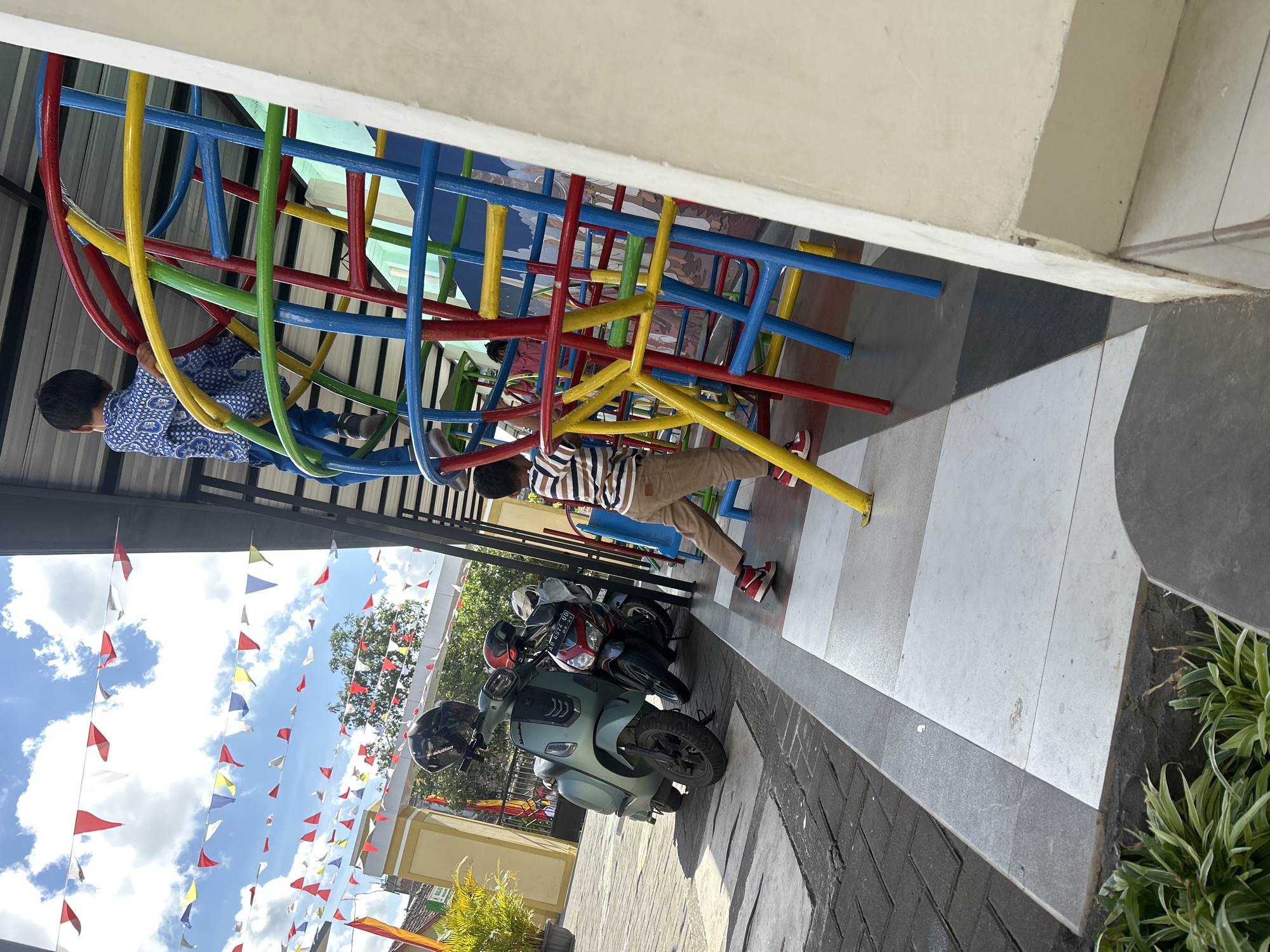 In the city of Wonosari, Indonesia, two young boys are engaged in activities on a brightly colored playground structure. The playground is a prominent feature, constructed with red, yellow, green, and blue metal poles forming a spherical frame. One boy, wearing a blue patterned shirt and blue pants, is positioned higher up on the structure, holding onto a yellow bar. The other boy, dressed in a striped shirt and brown pants, is standing on a lower section of the playground, looking towards the structure.
The playground is situated outdoors, under a covered walkway with a metal roof. In the background, a series of colorful triangular flags are strung across the sky, suggesting a festive occasion. A light green scooter with a black helmet and a red helmet rests on the paved ground to the left of the playground. Behind the scooter, a red motorcycle is parked. The ground is a mixture of gray and white tiles, with darker gray paving stones to the left. Lush green plants are visible in the foreground and in the background, adding to the outdoor setting. A yellow building with a red-tiled roof is partially visible behind the flags and trees. The sky is a bright blue with scattered white clouds.