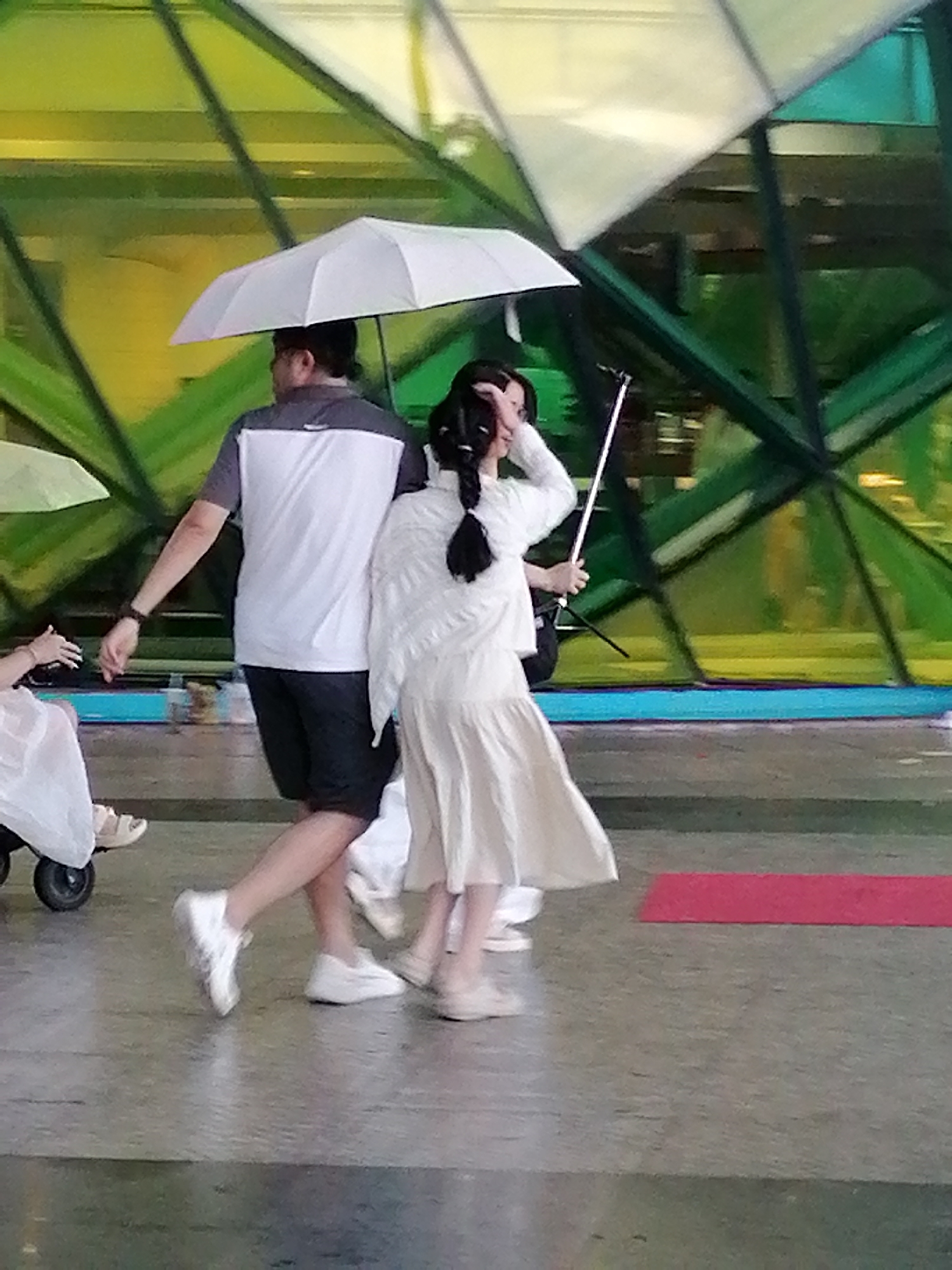 The image captures a candid street scene in Da Lat, Vietnam. A man wearing a gray and white t-shirt and black shorts, holding a white umbrella, walks alongside a woman in a flowing white dress. The woman is holding a selfie stick with a phone attached, seemingly capturing a photo or video. In the background, a colorful, angular structure with green and yellow panes suggests a modern architectural element, possibly a building or a public art installation. Other individuals are partially visible in the frame, including a person in a white outfit on a stroller-like apparatus to the left, and another person in a white dress to the far right. The ground appears to be paved, with a red mat laid out on the surface in the lower right quadrant. The lighting suggests it is daytime, and the presence of umbrellas indicates either a sunny or potentially rainy day. The overall atmosphere is casual and suggests people moving through an urban or public space.