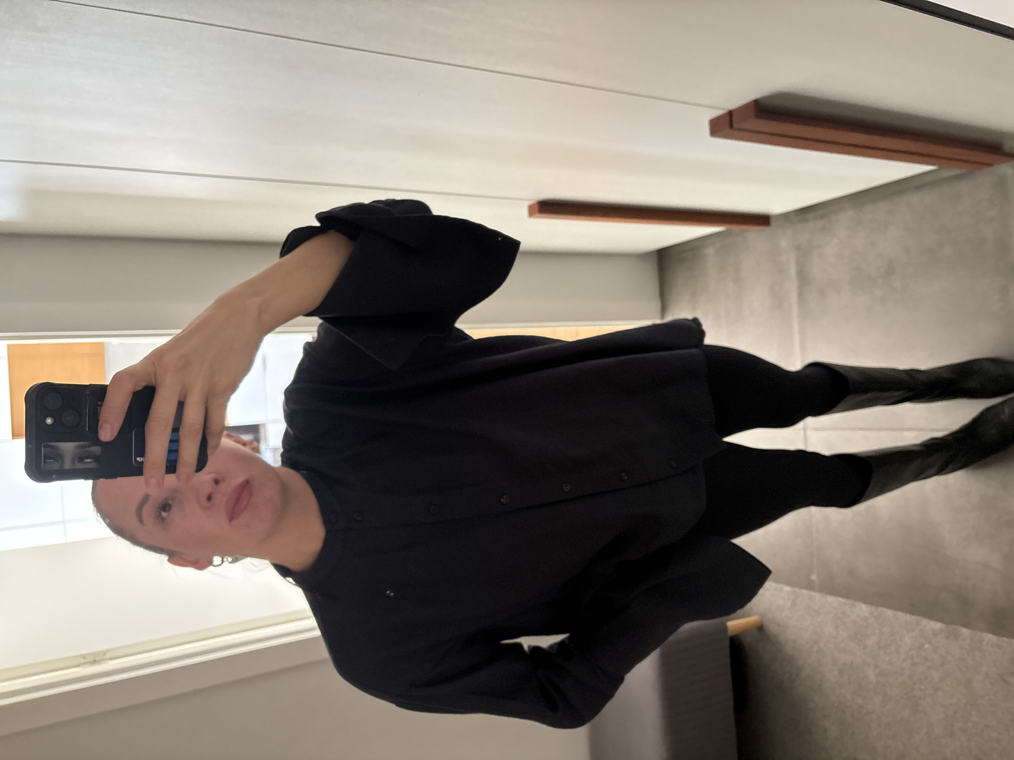 A person is taking a selfie in a room. The person is wearing a black outfit consisting of a collared, button-down shirt with wide sleeves, leggings, and boots. The phone being used to take the selfie is black. The room has a light-colored wall and a tiled floor. There is a dark-colored beam along the ceiling. Part of a gray seat is visible. The photo appears to have been taken in Chaikovsky, Russia.
