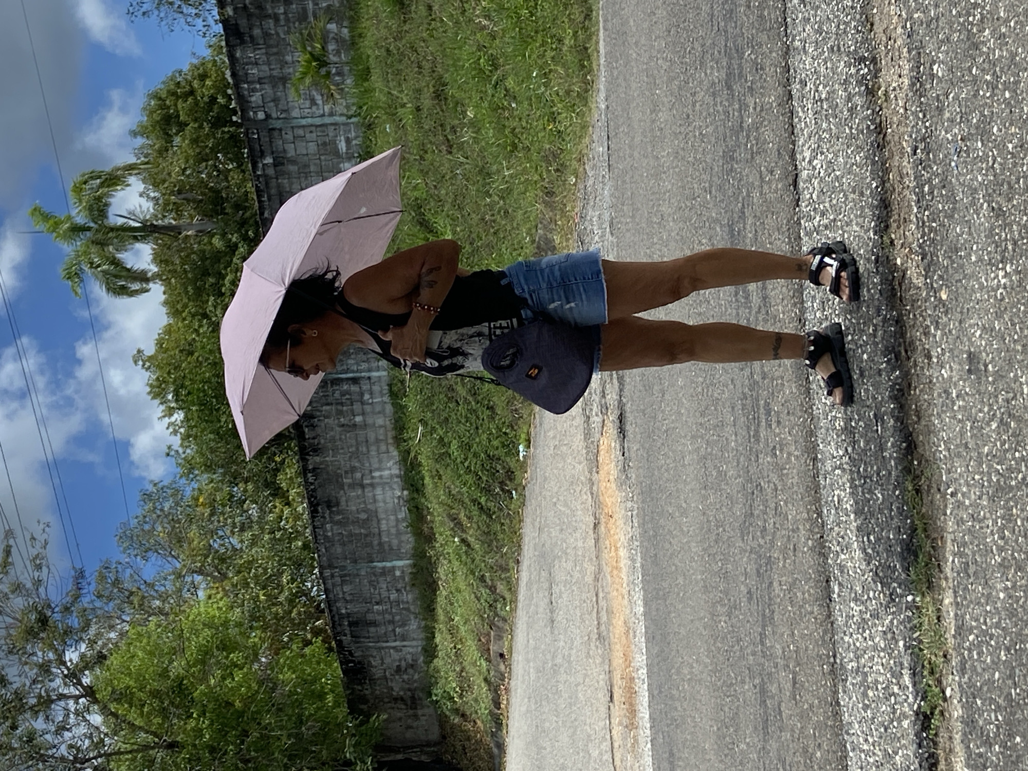 A person stands on a paved road, holding a light pink umbrella. The person is wearing a black tank top, a denim skirt, and black sandals. They also have a dark-colored bag slung over their shoulder. The background shows green vegetation, a concrete wall, a palm tree, and a partly cloudy sky. The location is likely in a tropical or subtropical area. Given the instructions, the image is set in the city of Rivero, Venezuela.