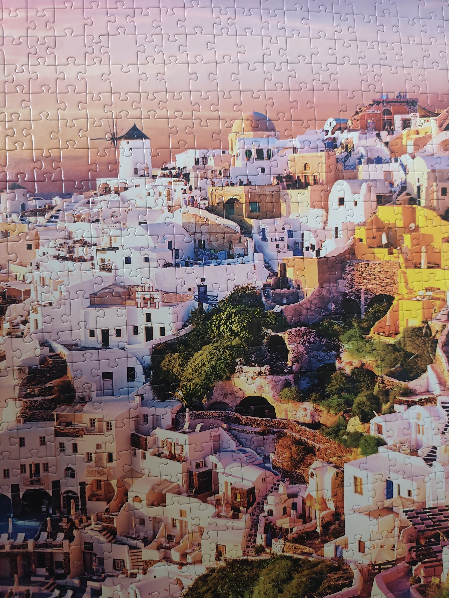 The image is of a partially completed jigsaw puzzle depicting the village of Oia in Santorini, Greece. The scene showcases the characteristic white-washed buildings cascading down the hillside, interspersed with domes, archways, and a windmill, all under a pink and orange hued sky. Green vegetation is also visible amidst the buildings. The composition is rendered as a puzzle, with the interlocking pieces visible throughout the image.