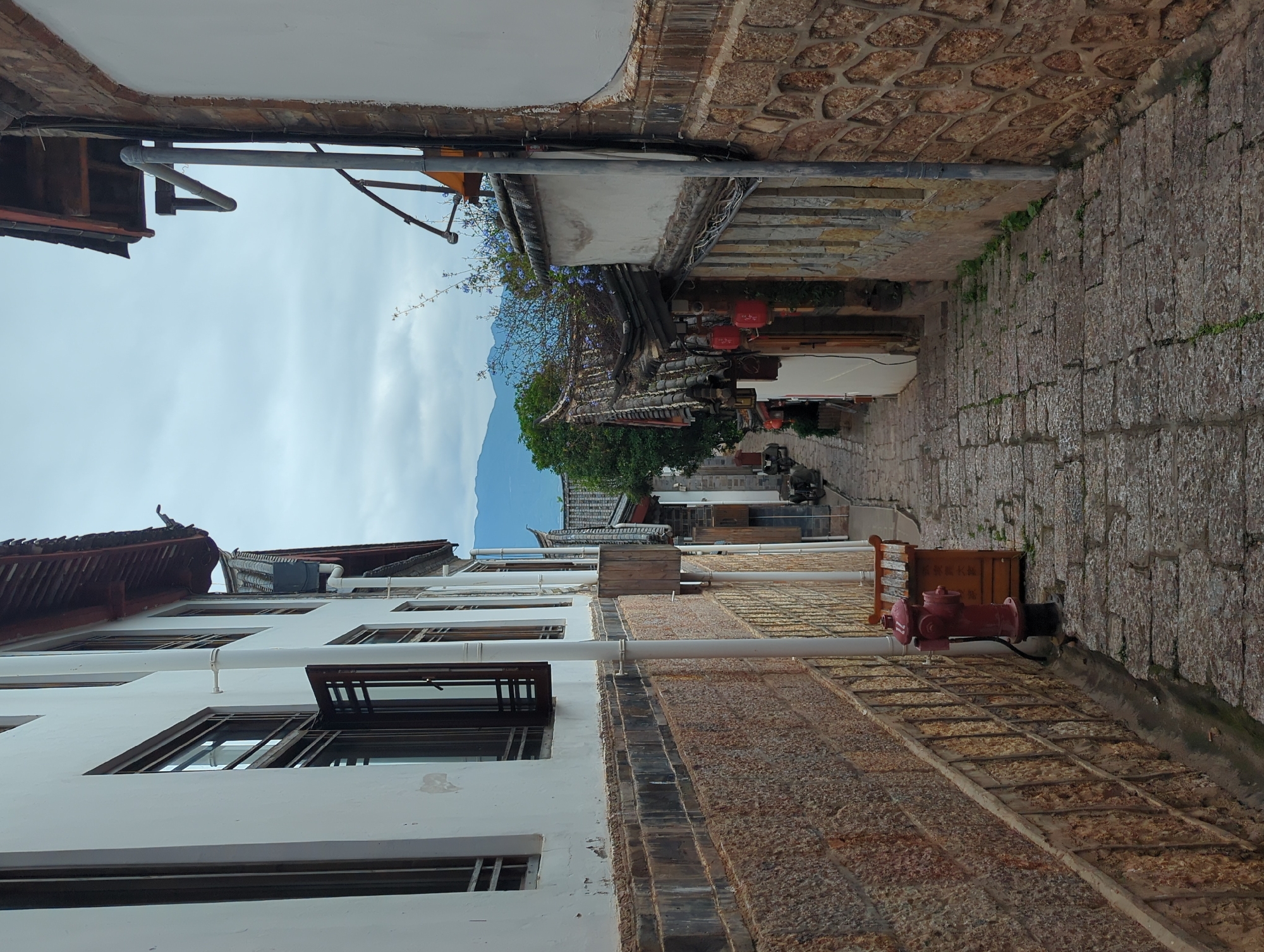 The image depicts a narrow, stone-paved alleyway in Gucheng District, China, flanked by traditional buildings. The buildings on either side feature white plastered walls, stone foundations, and dark tiled roofs with decorative eaves. The building on the left has several windows with dark wooden frames and one window is partially open. A white pipe runs vertically down the exterior of this building, attached to the stone wall. A red fire hydrant stands on the right side of the alleyway, next to a small wooden structure. The alleyway slopes downwards into the distance, where more buildings with red lanterns hanging from them are visible, along with some greenery. The sky is overcast with hints of mountains in the background.