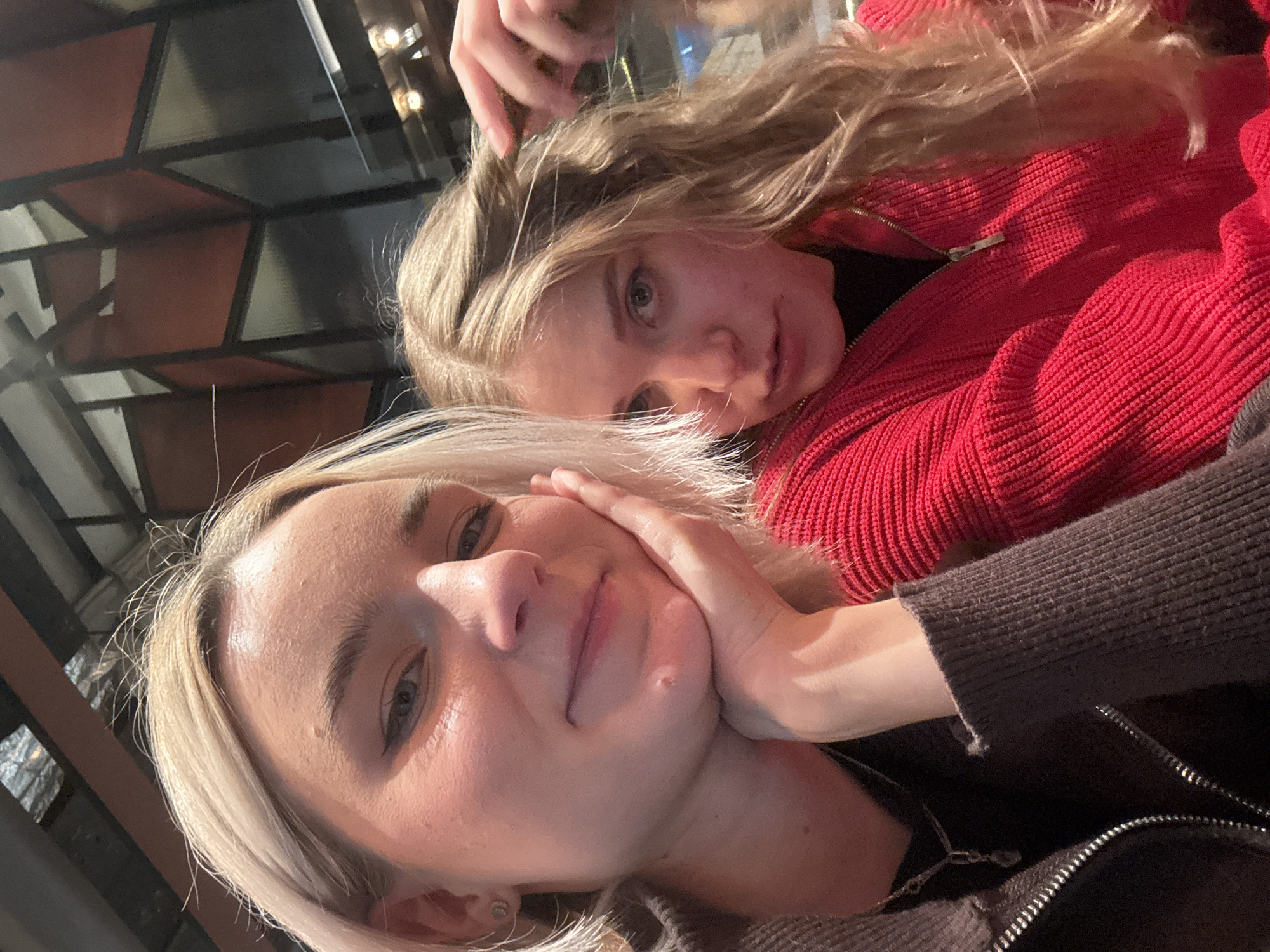 Two blonde women are in an indoor setting. One woman, wearing a brown zippered jacket, is closer to the camera with her face in her hand. The other woman is in the background wearing a red sweater. The location is in Unknown, Russia.