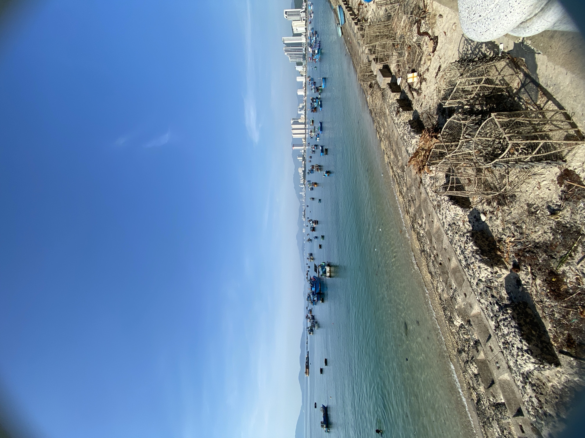 The image depicts a coastal scene in Bac Nha Trang Ward, Vietnam, featuring a calm sea dotted with numerous blue fishing boats. The water is clear and reflects the bright blue sky. In the background, a city skyline with tall buildings is visible, suggesting an urban coastal environment. The foreground shows a sandy shore with a concrete embankment. Several cylindrical, net-like structures, possibly fishing traps, are stacked on the embankment, along with debris such as dried leaves and branches. The overall atmosphere is serene, with the sea and sky dominating the composition.