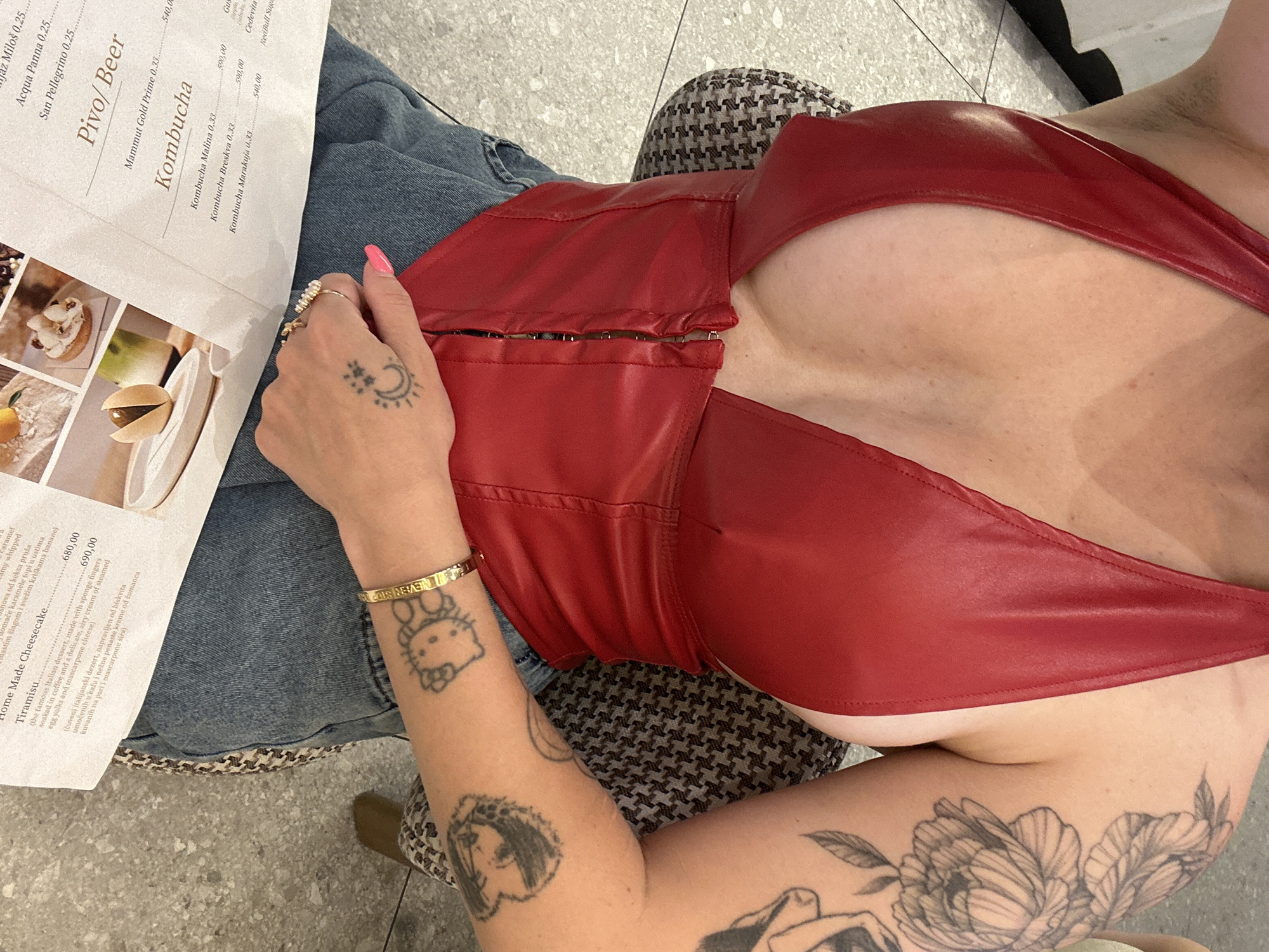 A close-up shot captures a person from the chest down, wearing a vibrant red halter top with a plunging neckline and a zipper closure. The individual has multiple tattoos on their left arm, including a Hello Kitty design and a floral pattern. They are also wearing rings on their right hand and a bracelet on their left wrist. The person is seated, with their hands resting on a menu or document that appears to be in a restaurant or cafe. The background shows a tiled floor and a patterned chair. The menu contains various food and drink items, with prices listed in what appears to be Serbian currency. Based on the text on the menu, specifically "Pivo/Beer" and the currency format, this image was likely taken in Belgrade, Serbia.