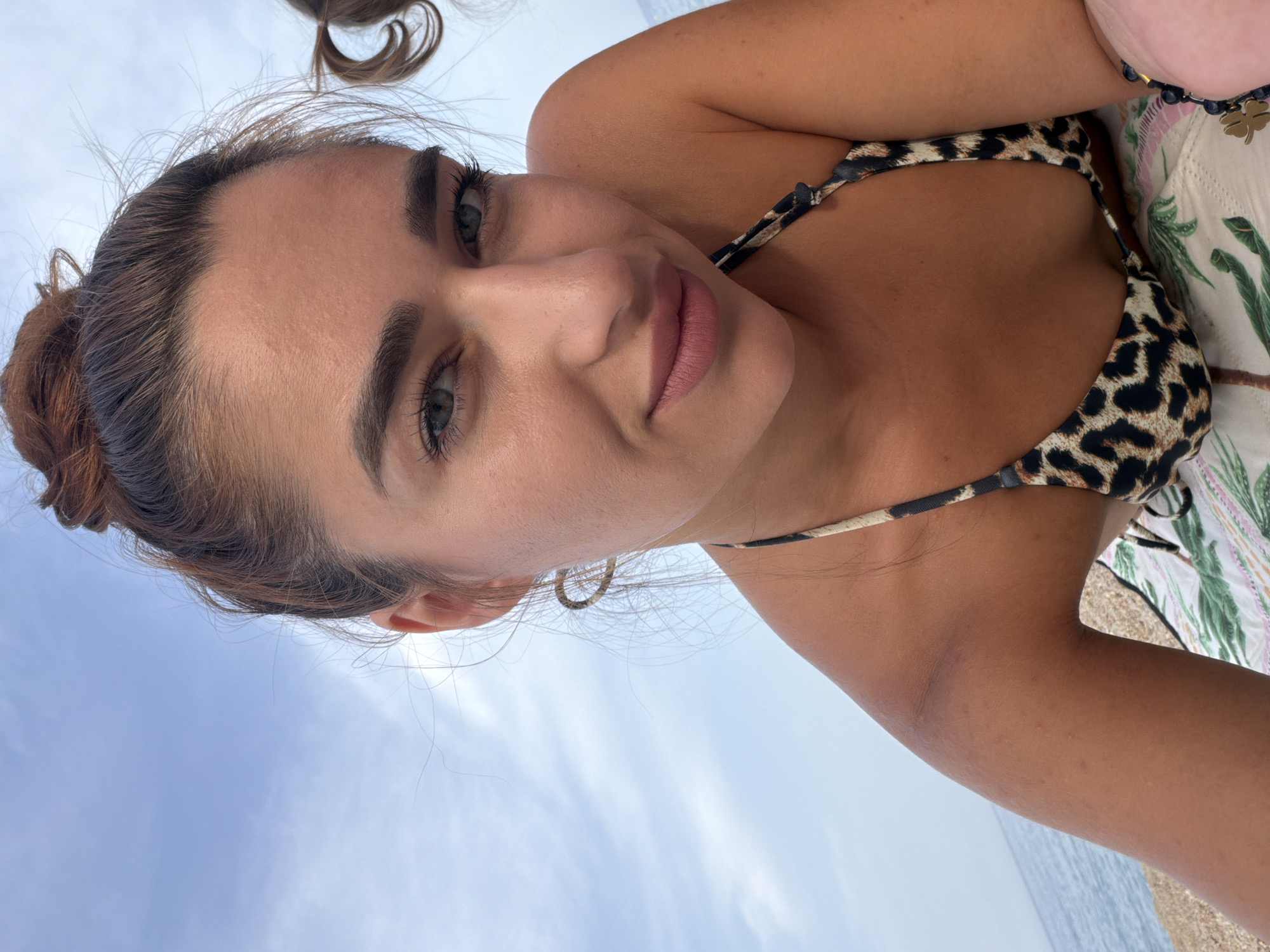 A woman with her hair in a bun and wearing a leopard print bikini top is lying down outdoors. She has on a bracelet with a clover charm. In the background, the sea and a cloudy sky are visible. The location is Platja D'aro, Spain.