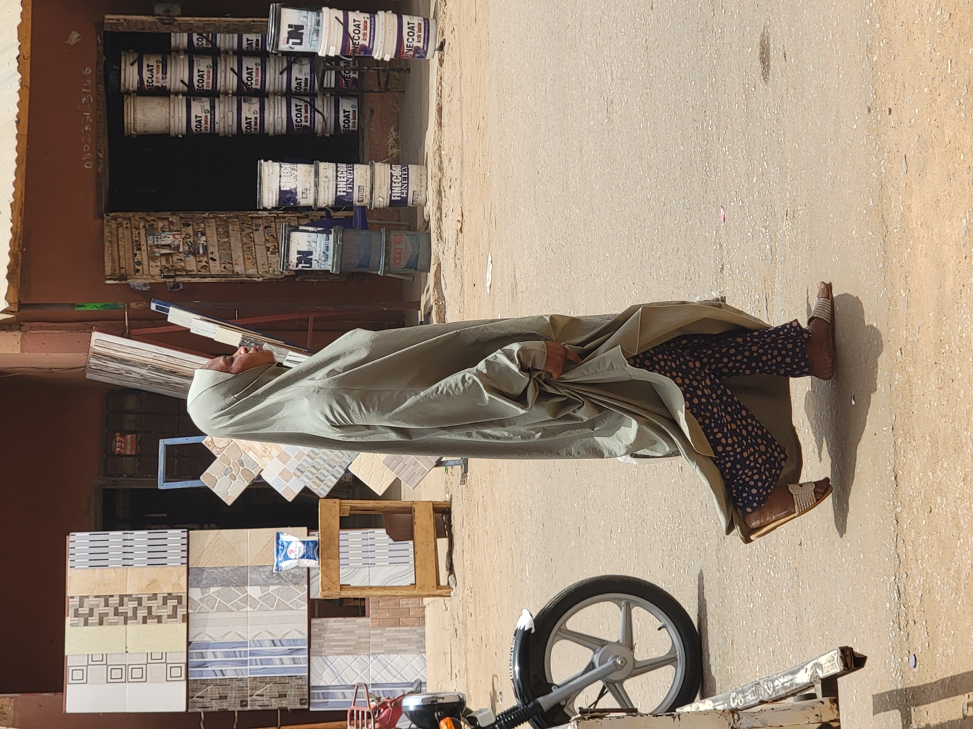 A man wearing sandals, dark spotted pants, and covered in a light green cloak is lying on a ladder next to a parked motorcycle near a building, likely in Maiburiji, Nigeria. The building has an awning and a storefront display of patterned tiles. Stacked cans of paint, some labelled "FINECOAT" and "ECOAT", are visible near an open section of the building, and a phone number "080253-3166" is painted on the wall.