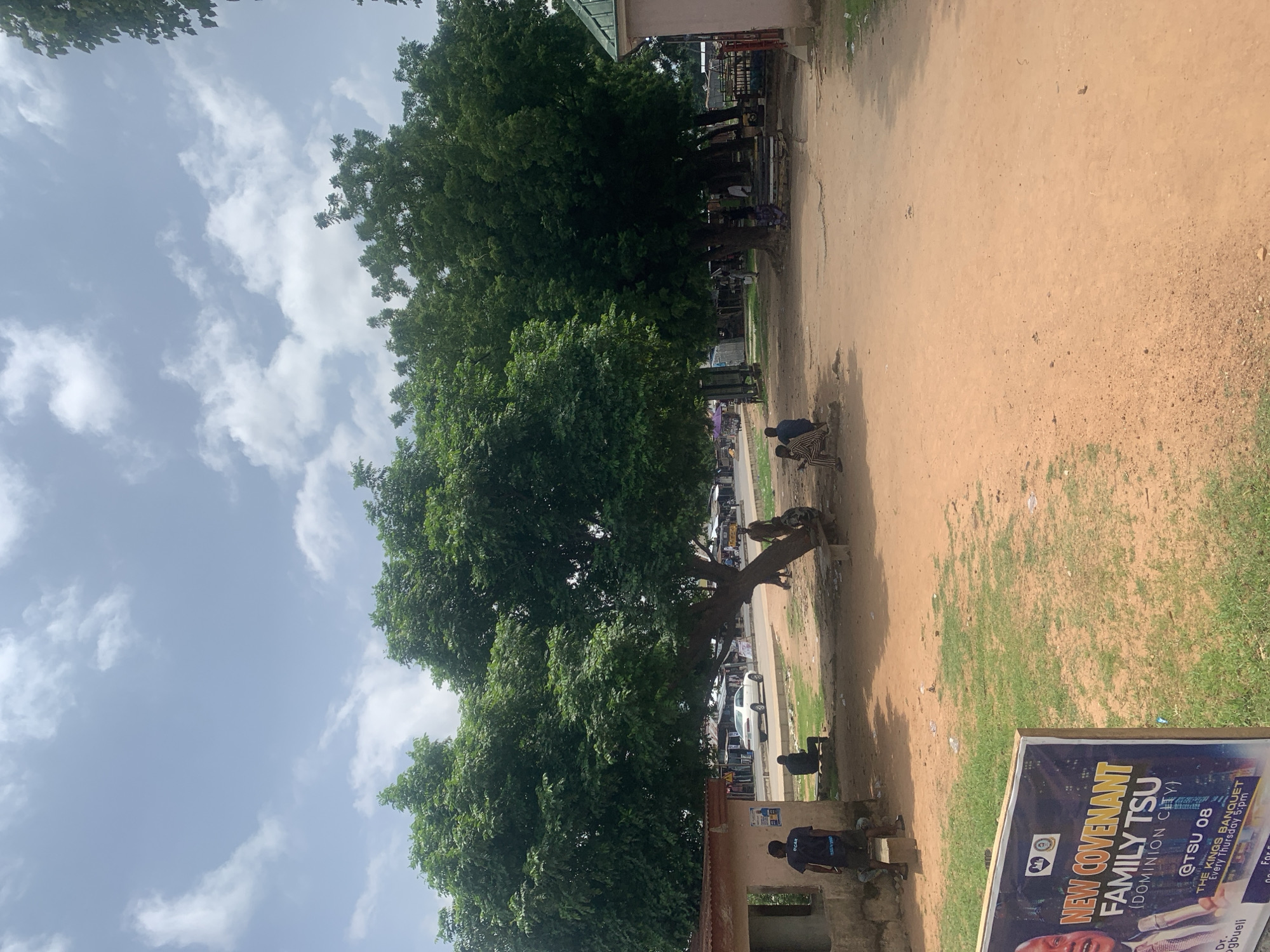 This is a vertical, eye-level, medium shot of a street scene in Jalingo, Nigeria. The foreground is dominated by a large tree with dense green foliage. The ground is a dirt path, with some patches of dry grass on the right side. In the mid-ground, there is a building with a green roof, and several people are visible sitting or standing near it. Behind the tree and building, a road is visible with several cars parked or driving by.
In the foreground on the right, a banner is partially visible. The visible text on the banner reads "NEW COVENANT FAMILY TSU (DOMINION CITY) @TSU 08 THE KINGS BANQUET Every Thursday 5:pm". Below this, there is an image of a man, and further text including "Dr. gbueli".
Several people are scattered throughout the scene, some sitting on benches or the ground, and others walking. Two individuals in the foreground on the right are wearing striped clothing. Another person in the mid-ground on the left is wearing a dark t-shirt with the text "CCAR" visible on the back.
The sky is mostly blue with scattered white clouds, suggesting it is daytime with fair weather. The overall scene depicts a typical day on a street in Jalingo, with people engaged in various activities under the shade of the tree and near the building.