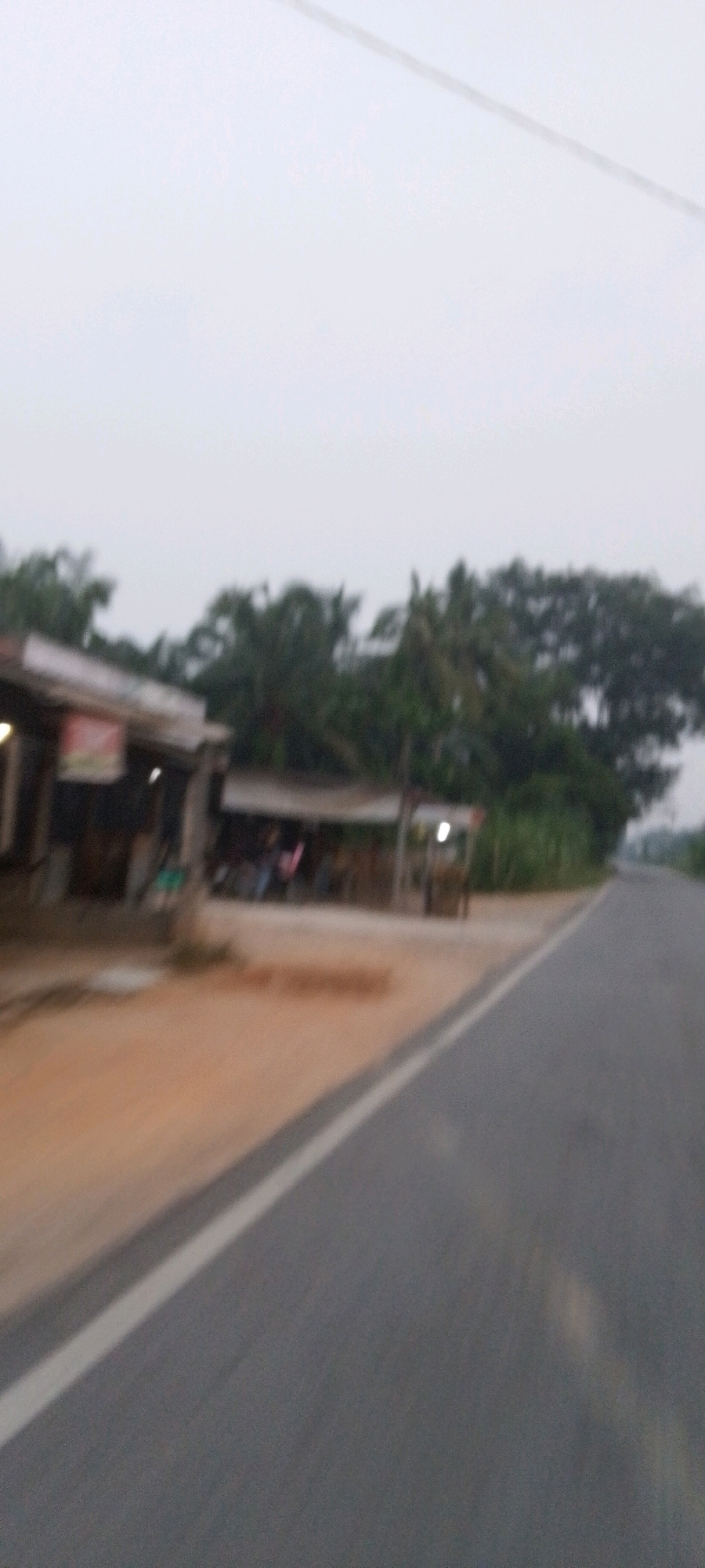 The image shows a blurred view of a road and some shops. The setting appears to be a rural or roadside area in Teluk Pulau Hulu, Indonesia. A gray asphalt road with a white line runs diagonally across the right side of the frame. On the left side, there are several small shops with open fronts, suggesting a market or small businesses. Some people are visible inside or near the shops, but they are indistinct due to the blur. Tropical trees and foliage form a backdrop behind the shops. The sky is overcast and a pale gray, indicating possibly dawn, dusk, or a cloudy day. The overall impression is a fleeting glimpse of a commercial area along a road in Indonesia.