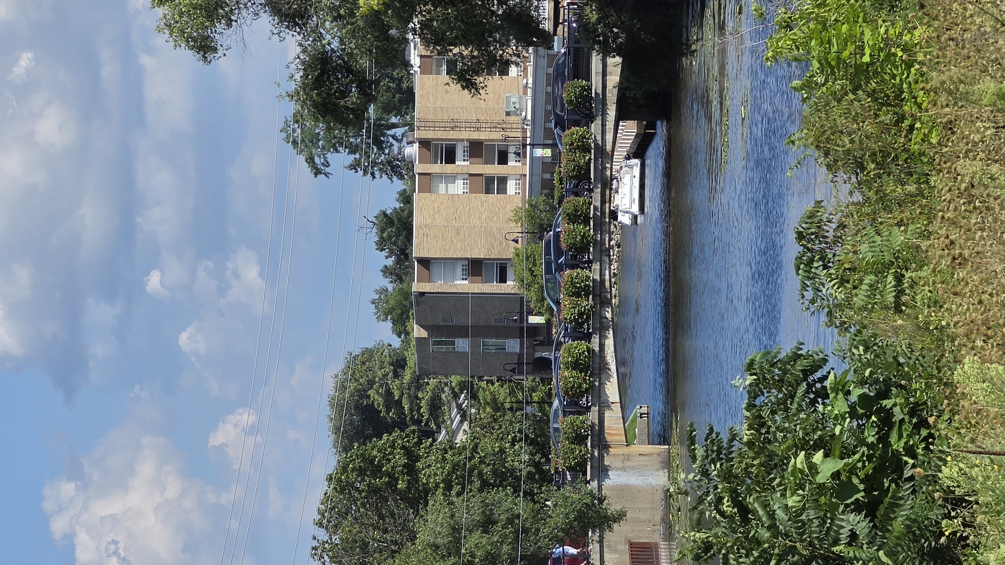 In McHenry, Illinois, United States of America, a body of water is visible, likely the Fox River, with a bridge crossing it. On the bridge, there are several parked cars, including a dark-colored sedan and a sport utility vehicle. A white boat is docked to the right of the bridge. Behind the bridge, a multi-story brick building with numerous windows stands, flanked by trees. The sky is blue with scattered white clouds. Green foliage covers the foreground and parts of the background, suggesting a natural environment within the urban setting. A few individuals are present on the left side of the bridge.