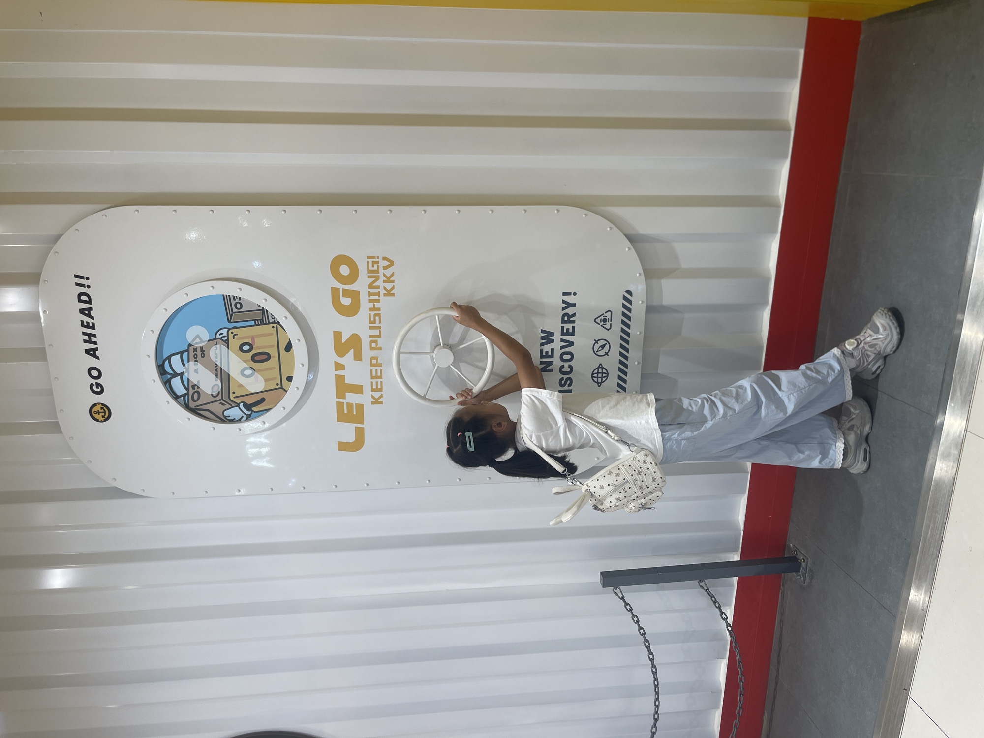 In an indoor environment, a young girl with dark hair in a ponytail, wearing a white t-shirt, light blue pants, and sneakers, stands facing a large white panel with a circular graphic and a steering wheel. She is holding the steering wheel with her right hand. The panel has text that reads "GO AHEAD!!" and "LET'S GO KEEP PUSHING! NEW DISCOVERY!". A graphic of a box character is visible through the circular graphic. The girl is carrying a white backpack adorned with a butterfly pattern. The background consists of a white corrugated wall and a yellow vertical section on the right. The floor is dark gray with a metal strip and a red baseboard. A chain barrier is visible to the left of the girl.