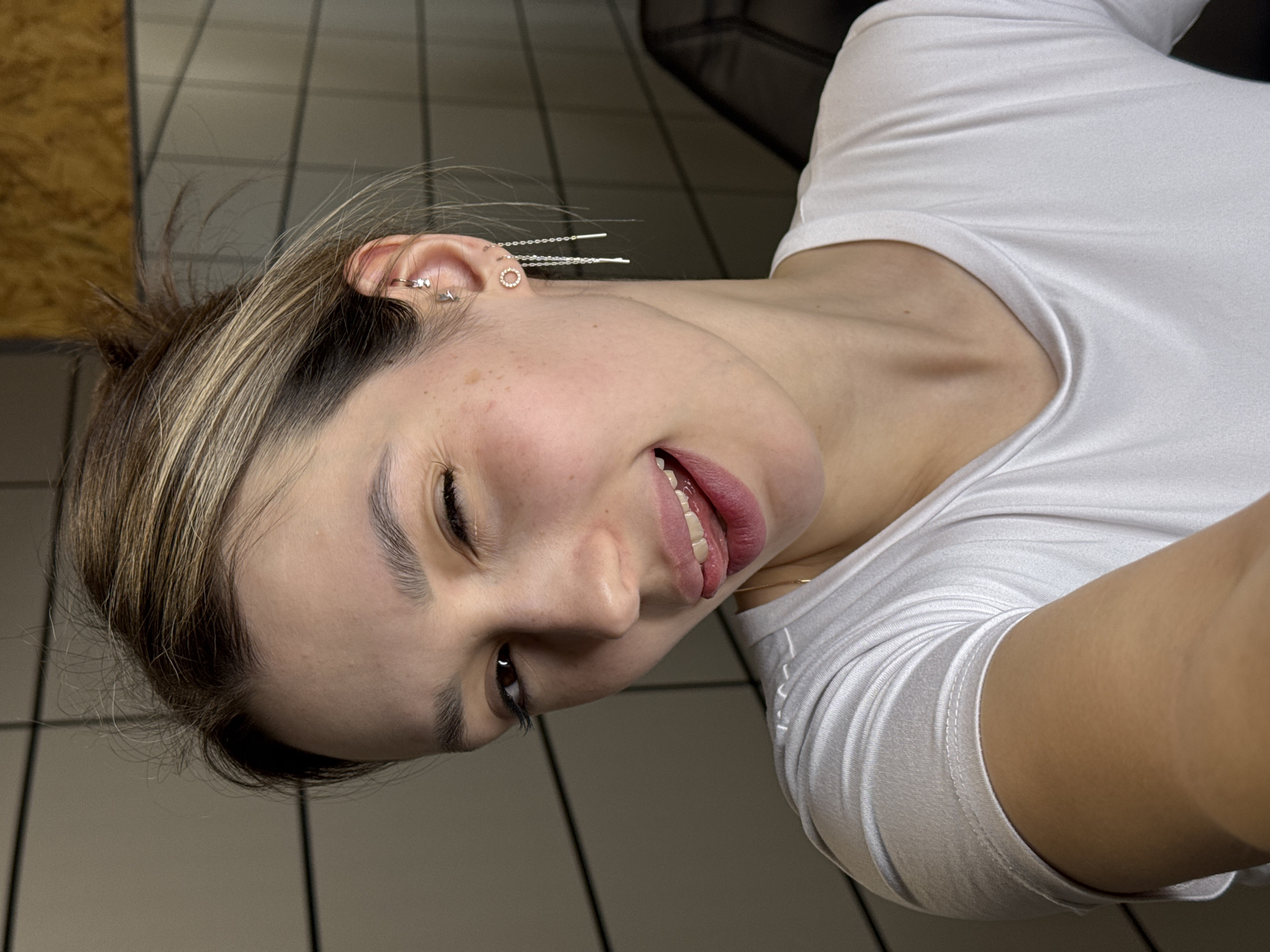A close-up, diagonal selfie captures a young person, likely female, with fair skin and light brown hair featuring blonde highlights. The person is wearing a white short-sleeved t-shirt. Their head is tilted significantly to the left, and they are smiling broadly with an open mouth, revealing teeth and a glimpse of their tongue, suggesting a playful or silly expression. Their right eye (viewer's left) is partially closed or squinting.
Visible details include several earrings on the right ear: a small hoop with a star charm, a circular stud, and two longer, chain-style earrings that thread through multiple piercings, featuring a bar at each end. A delicate golden chain is also visible around their neck. The person has visible freckles on their right cheek. Their eyebrows are dark and well-defined, and eyelashes appear long. The background shows light gray or beige square tiles with dark grout lines, suggesting an indoor setting, possibly a bathroom or a room with similar flooring or wall decor. There are also hints of wooden paneling in the upper left corner. The lighting appears to be artificial. No visible text is present in the image.
