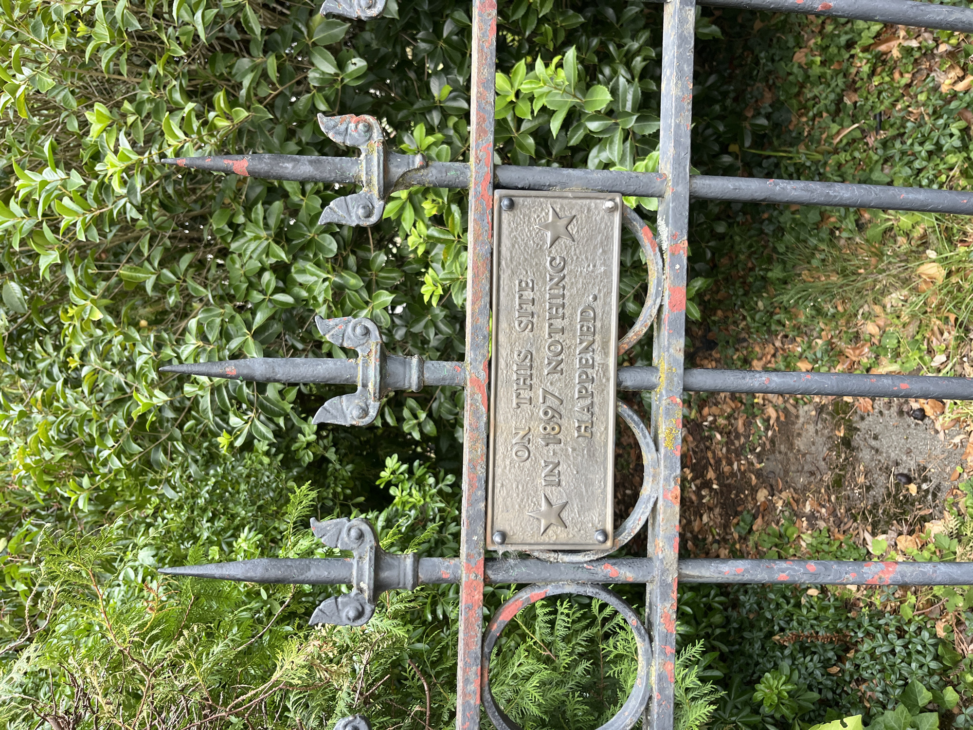 The image shows a close-up view of an ornate, dark grey metal gate with red paint chipping off, set against a backdrop of lush green foliage. Attached to the horizontal bar of the gate is a rectangular brass plaque with raised text and a star emblem. The plaque reads: "ON THIS SITE IN 1897 NOTHING HAPPENED." The gate features decorative spikes at the top and circular cutouts. The ground behind the gate is covered with fallen leaves and some patchy green ground cover. The overall impression is one of an aged, possibly historical, marker or monument situated within a garden or park setting. No people are visible in the image.