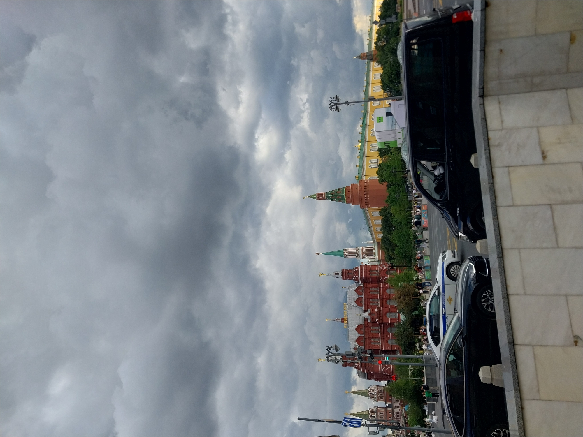 The image shows a street scene with several cars parked alongside a sidewalk. In the background, there are several historic buildings with red brick walls and green-topped towers, suggesting a location with significant architecture. The sky is heavily overcast with dark, dramatic clouds, indicating a cloudy day, possibly before or after a storm. The time of day is not precisely discernible due to the cloud cover, but the lighting suggests it is daytime.
On the street, a dark-colored van is parked closest to the viewer, followed by a white police car with markings that appear to be in Cyrillic script. The police car has the word "ПОЛИЦИЯ" visible on its side. Several other vehicles are partially visible further down the street. Pedestrians are walking on the sidewalk in the background, and trees are visible, adding to the urban landscape.
Notable details include the contrasting styles of the historic buildings and the modern vehicles. The starkness of the cloudy sky adds to the overall mood of the scene. The presence of a police car might suggest a public area or an event being monitored. There is some text visible on the white van in the background which is partially obscured by a tree, and the word "АПТЕКА" is visible on a green sign.