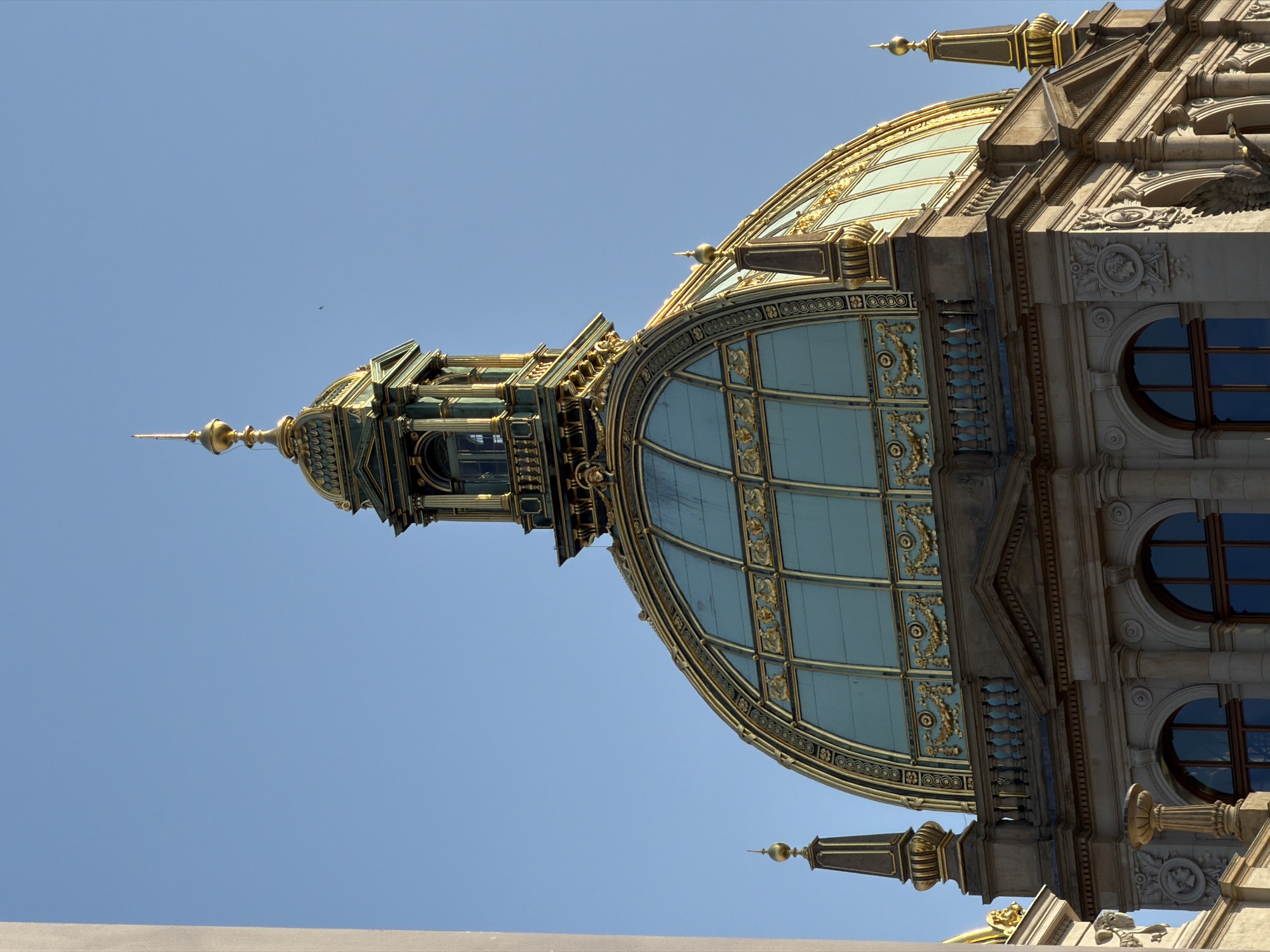 This is a low-angle, rotated shot of the ornate dome and part of the facade of a building against a clear blue sky. The building features a large, segmented glass dome with gold detailing, sitting atop a stone structure adorned with intricate carvings, classical columns, and arched windows. A gilded spire crowns the top of the dome structure. The overall impression is of grandeur and historical architecture. The lighting suggests it is daytime, likely sunny. No people or specific activities are visible. The crops provide closer views of the architectural details. Based on the location context, this is likely a building in Prague, Czechia.