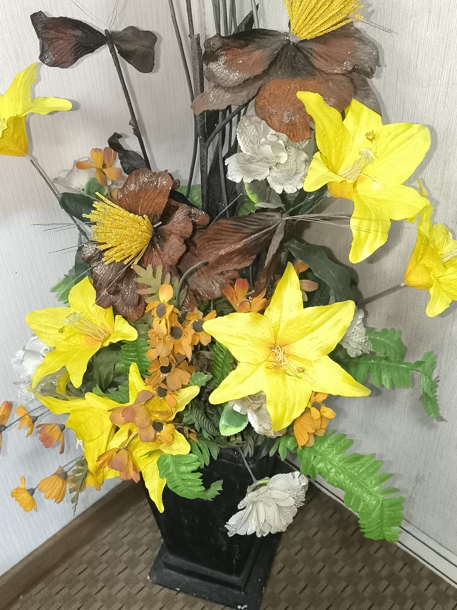 The image displays an artificial floral arrangement in a black vase. The arrangement features large, bright yellow lily-like flowers, smaller orange flowers, brown and dark brown ornamental leaves, and some green fern-like foliage. The flowers and leaves are positioned in a dense cluster, creating a visually striking display. The arrangement is placed against a textured, light-colored wall. The floor below the vase has a brown, tiled pattern. The image appears to be taken indoors, likely in a domestic or commercial setting. There is no indication of the specific location within Kalabbirang, Indonesia, based on the visual information provided.
