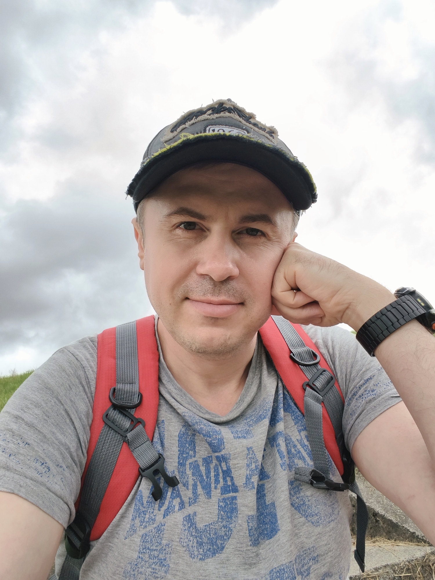 A man is taking a selfie outdoors. He is wearing a grey t-shirt with a blue graphic design and a black baseball cap. He has a red backpack on. His right arm is propped up on his chin and he is looking directly at the camera. The background is a cloudy sky and a green grassy hill. There is a concrete ledge in the lower right corner of the image. This image was taken in Tczew, Poland.