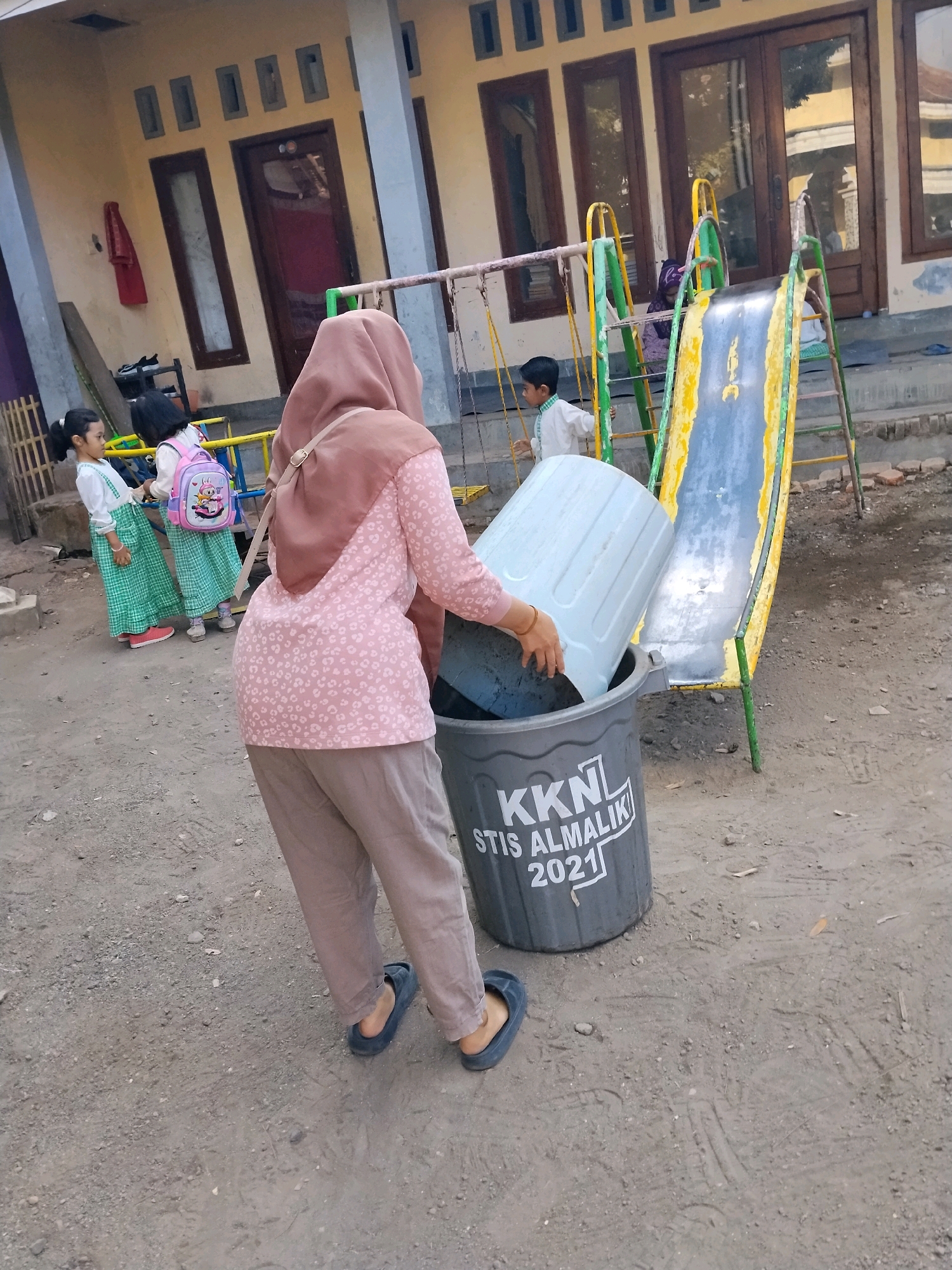 In Panggungan, Indonesia, a woman wearing a pink headscarf and a pink floral shirt is seen from behind, attempting to place a large white plastic bin into a grey trash can. The trash can, which is labeled "KKN STIS ALMALIK 2021," is situated on a dirt ground near a playground with a yellow and blue slide. Two young girls in green checkered dresses and white tops stand to the left, one with a pink backpack. A young boy in a white shirt stands on the playground structure. The background shows a building with beige walls and dark brown doors and windows. The overall environment appears to be an outdoor area, possibly a school or community center, with a mix of play equipment and a residential building.