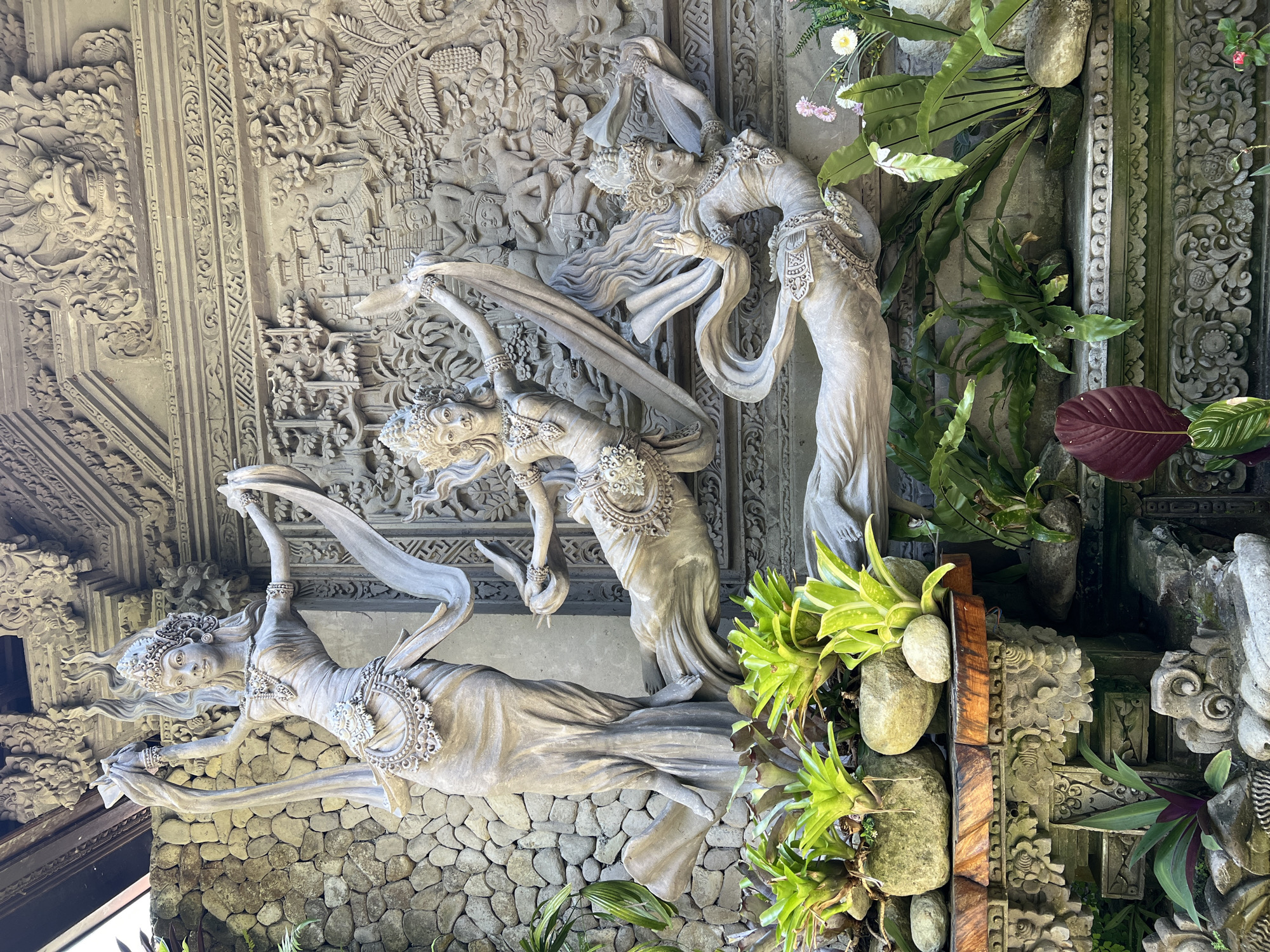 The image shows a wall in Kedewatan, Indonesia, adorned with sculptures. Three stone-colored figures of dancing women are positioned against a backdrop of stone carvings depicting a scene with multiple figures. The figures are wearing ornamented cloths, jewels and elaborate headdresses, and are frozen in fluid motions of dance. The background wall is decorated with carvings of figures and foliage. The base of the wall is constructed from stacked stones. The side of the display is adorned with assorted plants and stones.