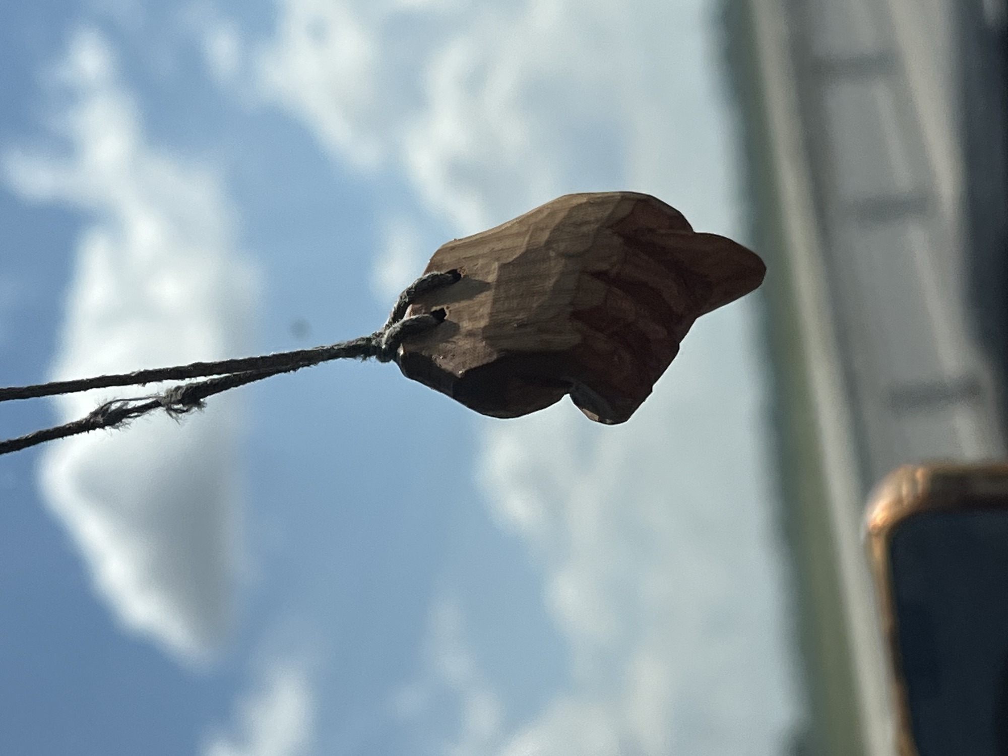 A close-up, horizontal shot captures a small, carved wooden object resembling a hand or fist, suspended by a frayed rope against a backdrop of a partly cloudy blue sky. The wooden carving is dark brown and appears textured with visible tool marks. It is attached to two strands of dark, weathered rope. The rope extends to the left and appears to be attached to something out of frame. The sky is bright, suggesting daylight, with fluffy white clouds scattered across the blue expanse. In the far right of the frame, a blurry vertical element with a golden-brown edge is visible, possibly the corner of a window or frame. The lighting and sky suggest it is daytime, likely a sunny or partly sunny day. There is no visible text. The setting appears to be outdoors with a view of the sky. The overall impression is simple and somewhat rustic, focusing on the natural textures of the wood and rope against the vastness of the sky.