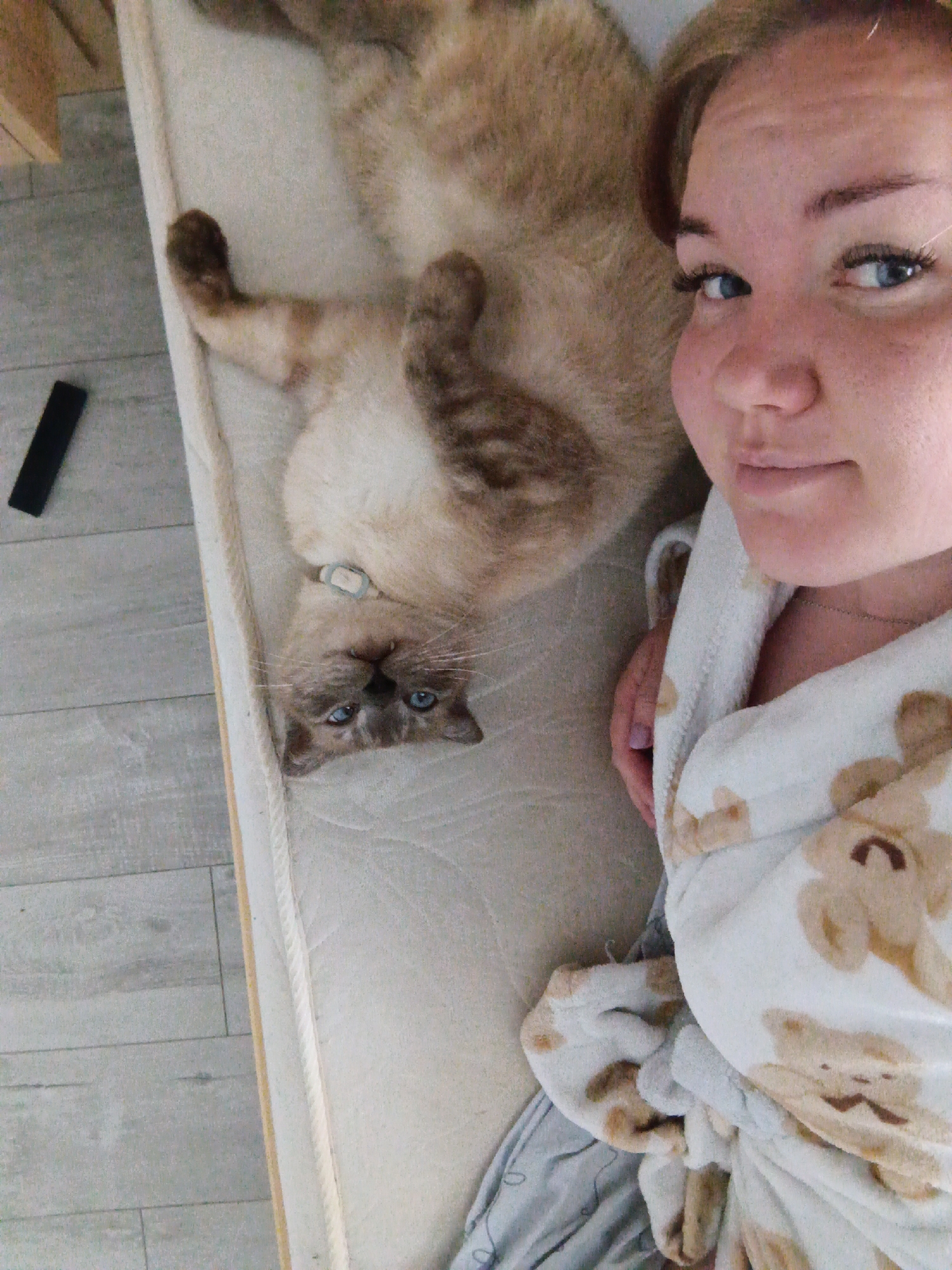 A woman with fair skin and blue eyes is lying on a mattress next to a beige and brown cat with blue eyes. The woman is wearing a white robe with teddy bear patterns. The cat is lying on its back, facing the camera. A black object is visible on the floor next to the mattress. The location is Фоки, Russia.
