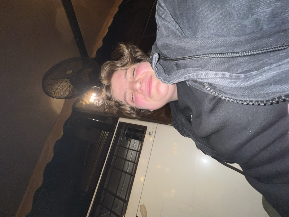 A person smiles at the camera while wearing a dark jacket. The background is poorly lit. An electric fan and a white box with a dark grate are visible behind the person. The photo is taken at an odd angle. The person has curly hair and appears to be indoors.
