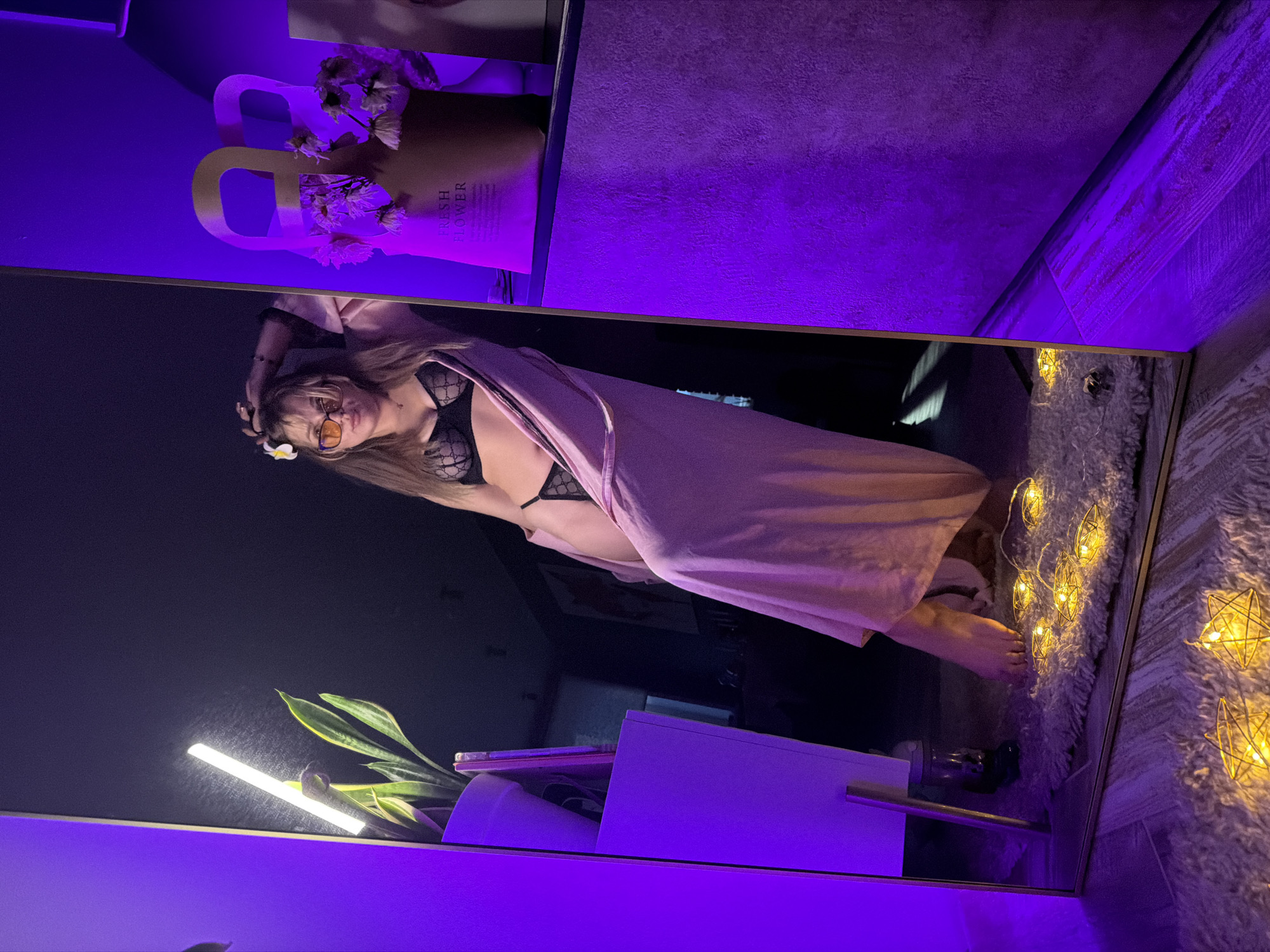 A reflection in a mirror shows a woman with long hair wearing a black lace bra and a pink robe. She is standing on a fluffy white rug with star-shaped string lights. To the left of the reflection are a plant and a white container with purple accents. Above the mirror, there is a flower arrangement in a brown paper bag with handles. The scene is lit with purple and blue lighting. The location is Большая Шильна, Russia.