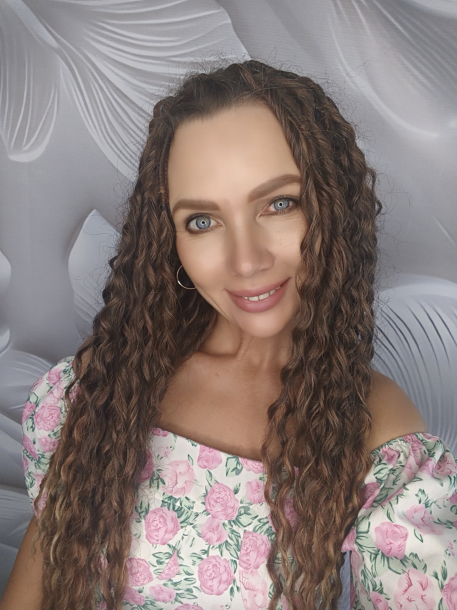 A woman with long, curly brown hair and blue eyes smiles at the camera. She is wearing a floral off-the-shoulder top with pink roses and green leaves. She has a hoop earring in her visible ear. The background is a light gray with a white textured pattern.