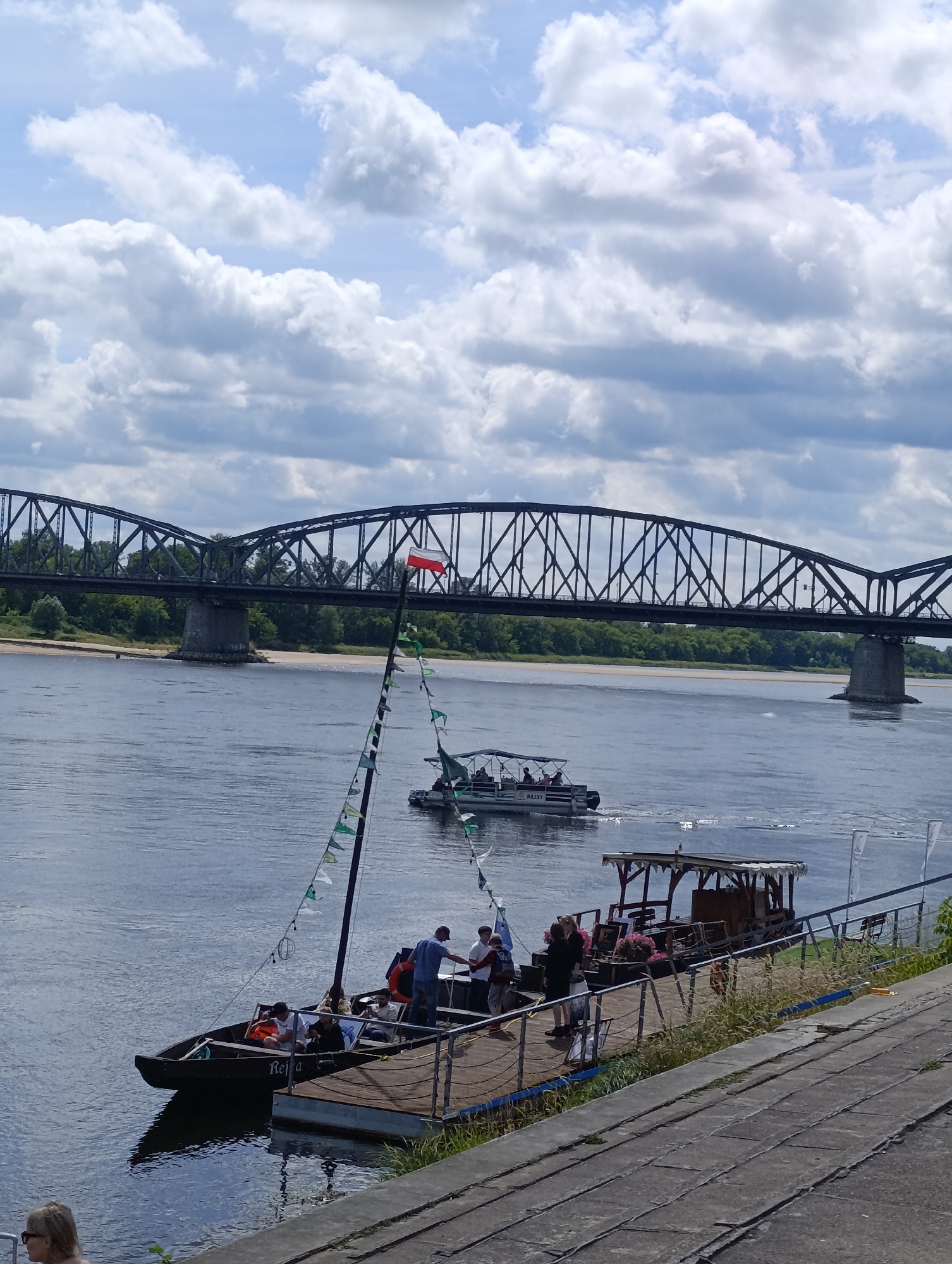 The image shows a view of the Vistula River in Toruń, Poland, with a large, dark metal truss bridge spanning the river in the background. In the foreground, a small wooden boat with a sail is docked at a wooden pier. Several people are on the boat and the pier. The boat has a red and white flag at the top of its mast, and a string of small green and white flags hangs from the mast. A second, larger boat, a pontoon boat, is visible on the river in the middle ground, also with several people on board. The river is wide and calm, reflecting the cloudy sky. The sky is filled with large, fluffy white clouds against a blue background. The far bank of the river is lined with trees. The pier appears to be made of wood planks, with some grassy vegetation growing around it. A woman with blonde hair is visible on the far left of the image, looking to her left.