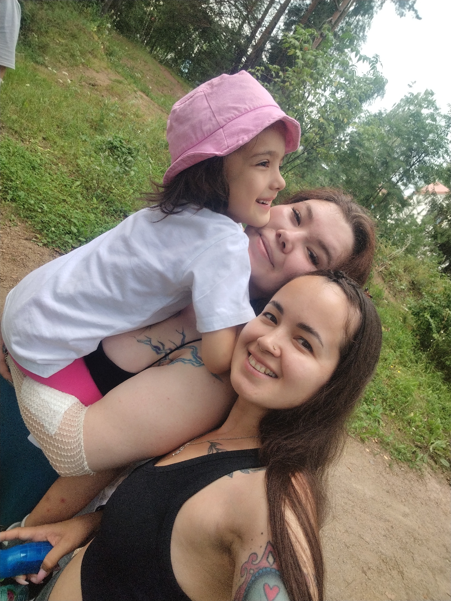 Three individuals, two women and a child, are positioned in a close-up, outdoor photograph. The foreground features the face of a woman with dark hair, wearing a black tank top and a nose ring, smiling broadly. Behind her, another woman with dark hair and a bright pink hat is partially visible, looking upwards. Nestled between them is a young child wearing a white t-shirt and the pink hat, smiling and looking to the side. The background consists of green foliage and trees, suggesting a natural, park-like setting. The ground appears to be a dirt path. The lighting indicates it is daytime, likely overcast. No specific landmarks or identifiable features of Noviy, Russia are present in the image.