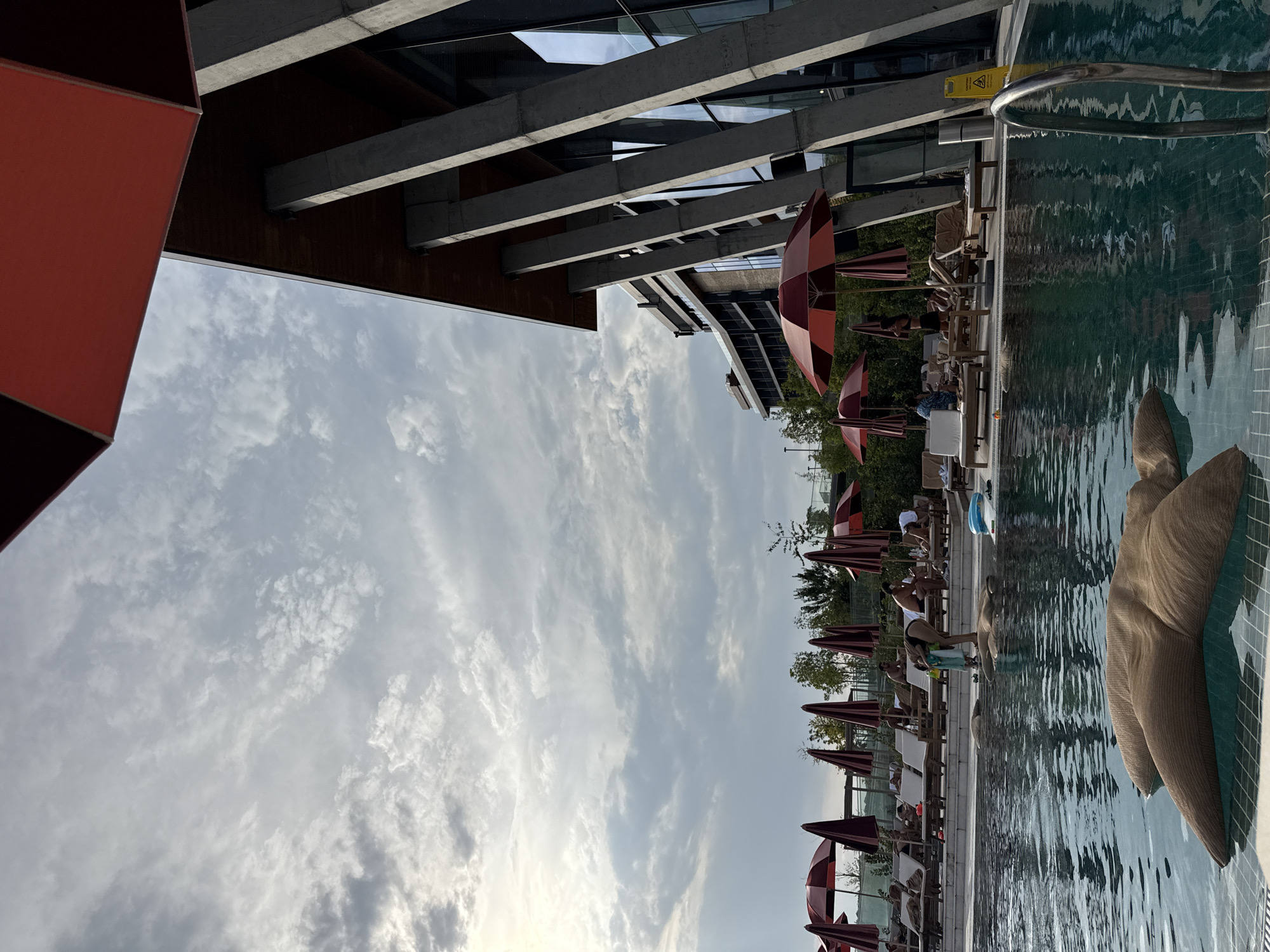 The image depicts a contemporary outdoor swimming pool area with a modern building in the background. The pool has clear green water, with a large, textured, star-shaped float in the foreground. A silver ladder leads into the pool from the right. Beyond the pool, lounge chairs with umbrellas are arranged, and several people are relaxing or walking. The building features a striking architectural design with large concrete pillars and glass windows. The sky is cloudy, with patches of sunlight breaking through. The scene suggests a resort or hotel setting. The city of Sighnaghi, Georgia, is not directly represented in the visual elements of the image.