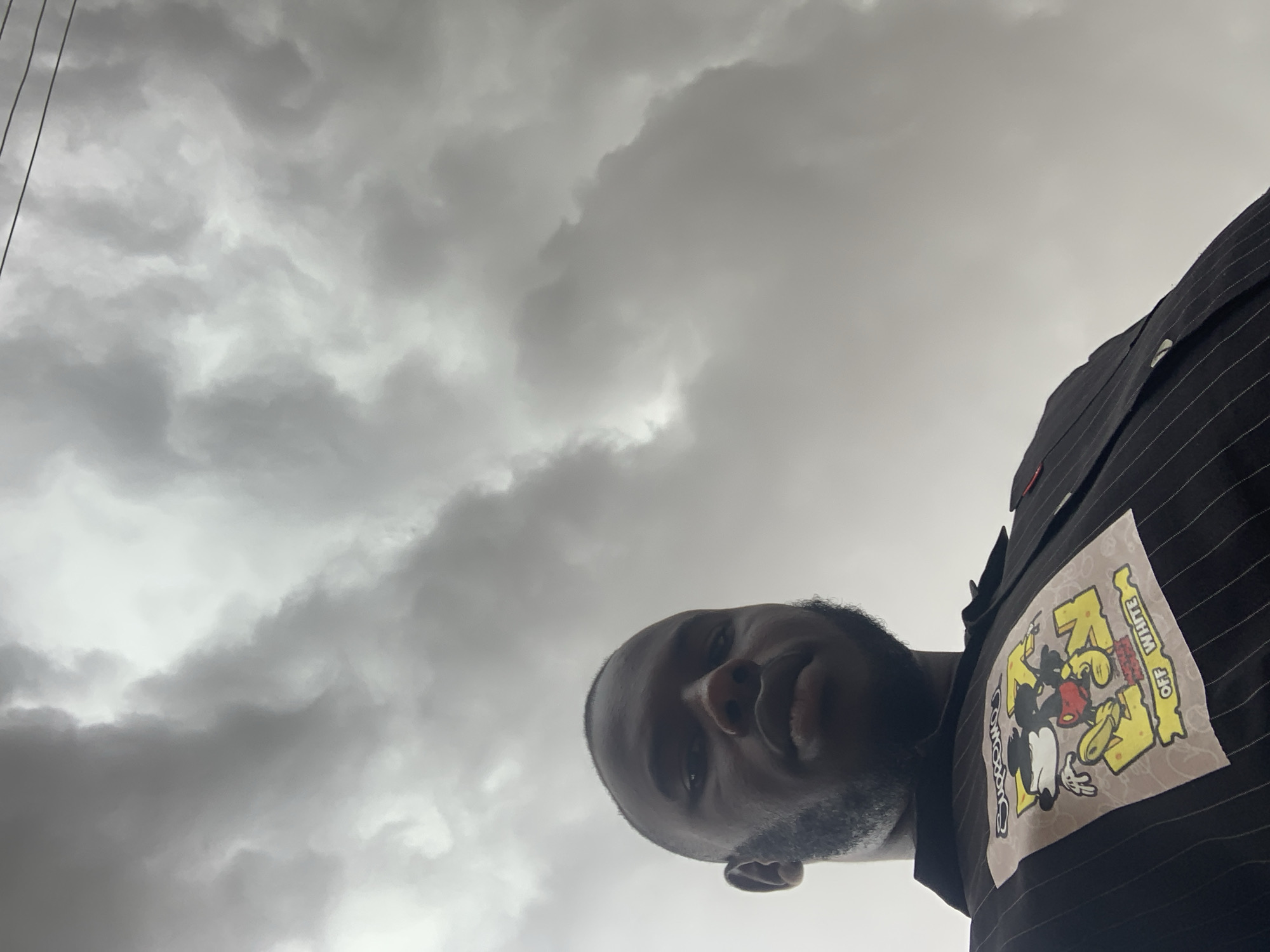 A low-angle shot captures an adult man against a heavily overcast sky in Jos, Nigeria. The man, with dark skin and a beard, is positioned in the lower right portion of the frame, looking downwards towards the camera. He is wearing a dark shirt or jacket, possibly pinstriped, which features a prominent square patch or print. This graphic depicts a stylized Mickey Mouse character with large yellow letters and shapes around it. Clearly visible text on the patch includes "OFF WHITE," oriented vertically, and fragmented text that appears to be "COMICS."
The sky dominates the upper and left areas of the image, filled with dense, dark grey clouds that suggest heavily overcast weather or an approaching storm. Thin, dark lines, resembling power lines or cables, are visible in the upper left corner against the cloudy backdrop. The lighting is diffused, indicating daytime. The man's expression is neutral, and the upward perspective creates a dynamic and expansive feel, contrasting the human subject with the vast, cloudy sky.