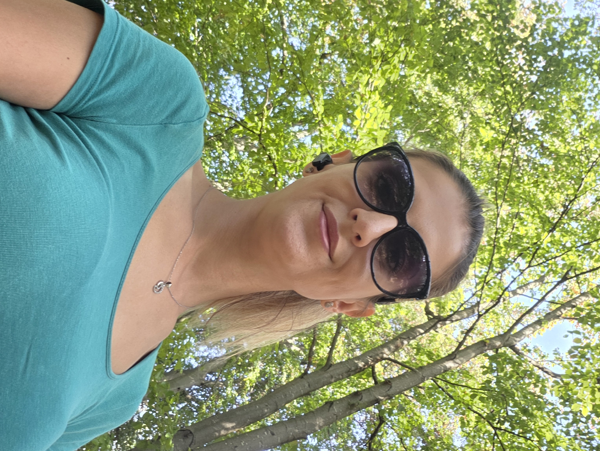 A low-angle, medium shot shows a woman from the chest up, looking at the camera. She has blonde hair pulled back and is wearing large, dark sunglasses, a teal t-shirt, and a silver necklace with a small pendant. She has a wireless earbud in her right ear. Behind her, the shot is dominated by the bright green leaves and brown trunks of trees, with a glimpse of the sky between the branches. The lighting suggests it is daytime and sunny. There is no indication of the image being taken in Munich, Germany.