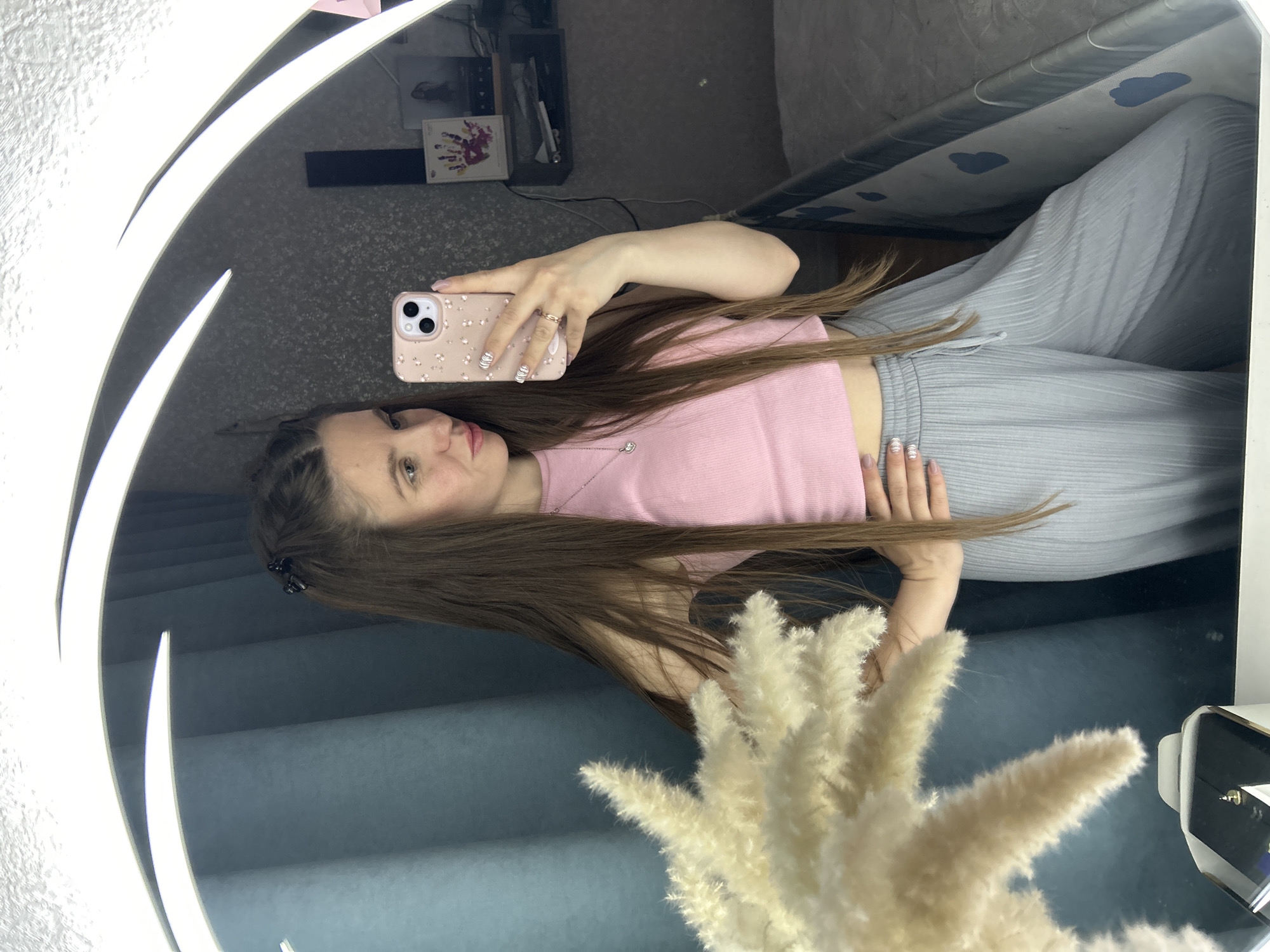 A woman is taking a selfie in a round mirror. She is wearing a pink top and grey pants. Her hair is long and brown. She is holding a pink phone with a decorative case. The background includes a grey wall, a shelf with items, and a blanket with a cloud pattern. A decorative plant is partially obscuring the view. The location is potentially Klin, Russia, but there's no verifiable data to prove this from the photo.