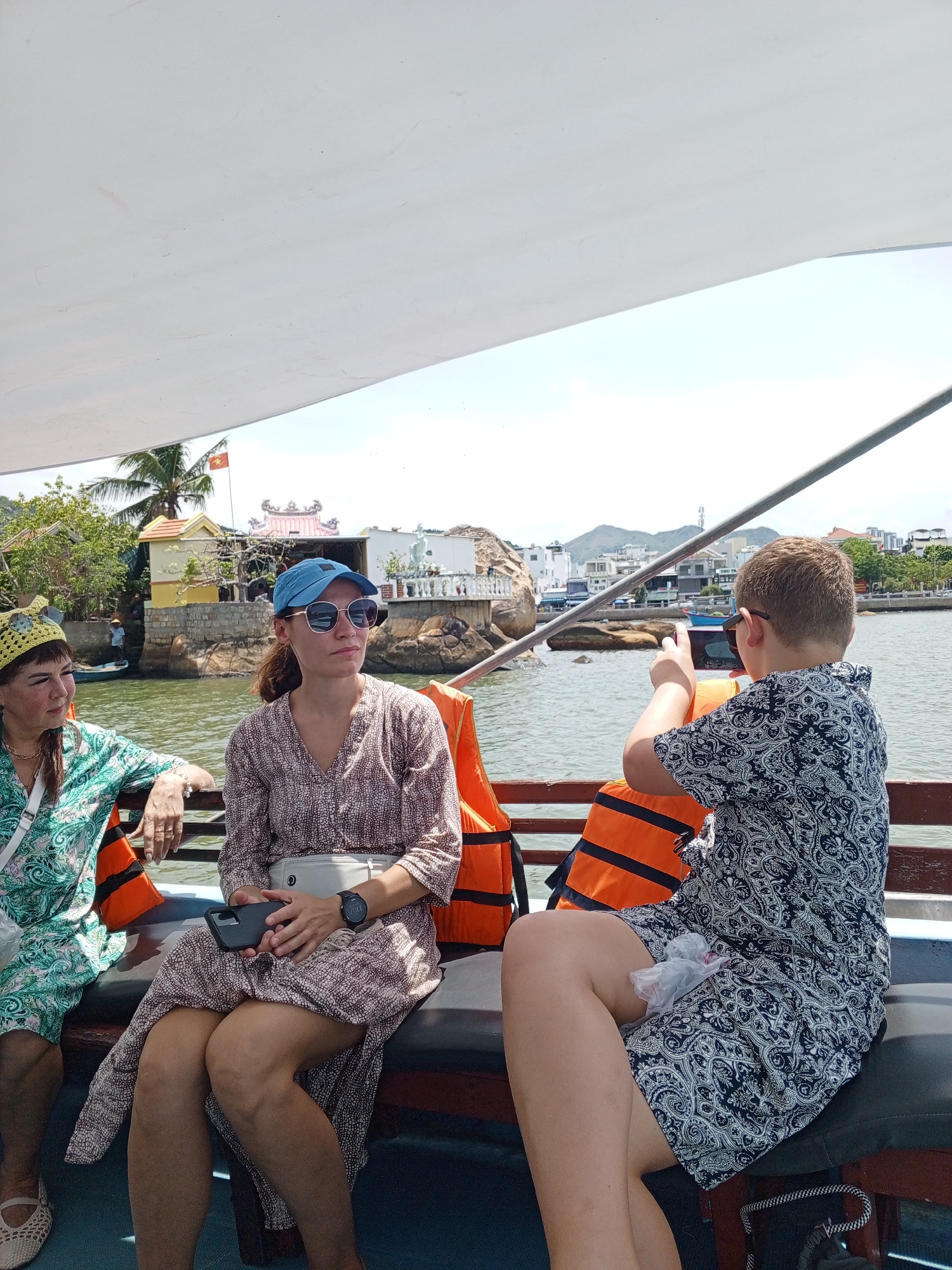 Three individuals are seated on a boat's bench under a white canopy. On the left, a woman wearing a yellow crocheted hat and a green patterned dress is looking to her left. In the center, a woman wearing a blue baseball cap, sunglasses, and a patterned dress is looking to her right and holding a black smartphone. To the right, a young person wearing a patterned shirt is turned away from the camera, using a smartphone to take a picture of the scenery. Two orange life vests are visible behind the seated individuals. The background shows a body of water with buildings, palm trees, and hills in the distance. The scene appears to be in Bac Nha Trang Ward, Vietnam, given the presence of a Vietnamese flag on a building and the distinctive architecture.