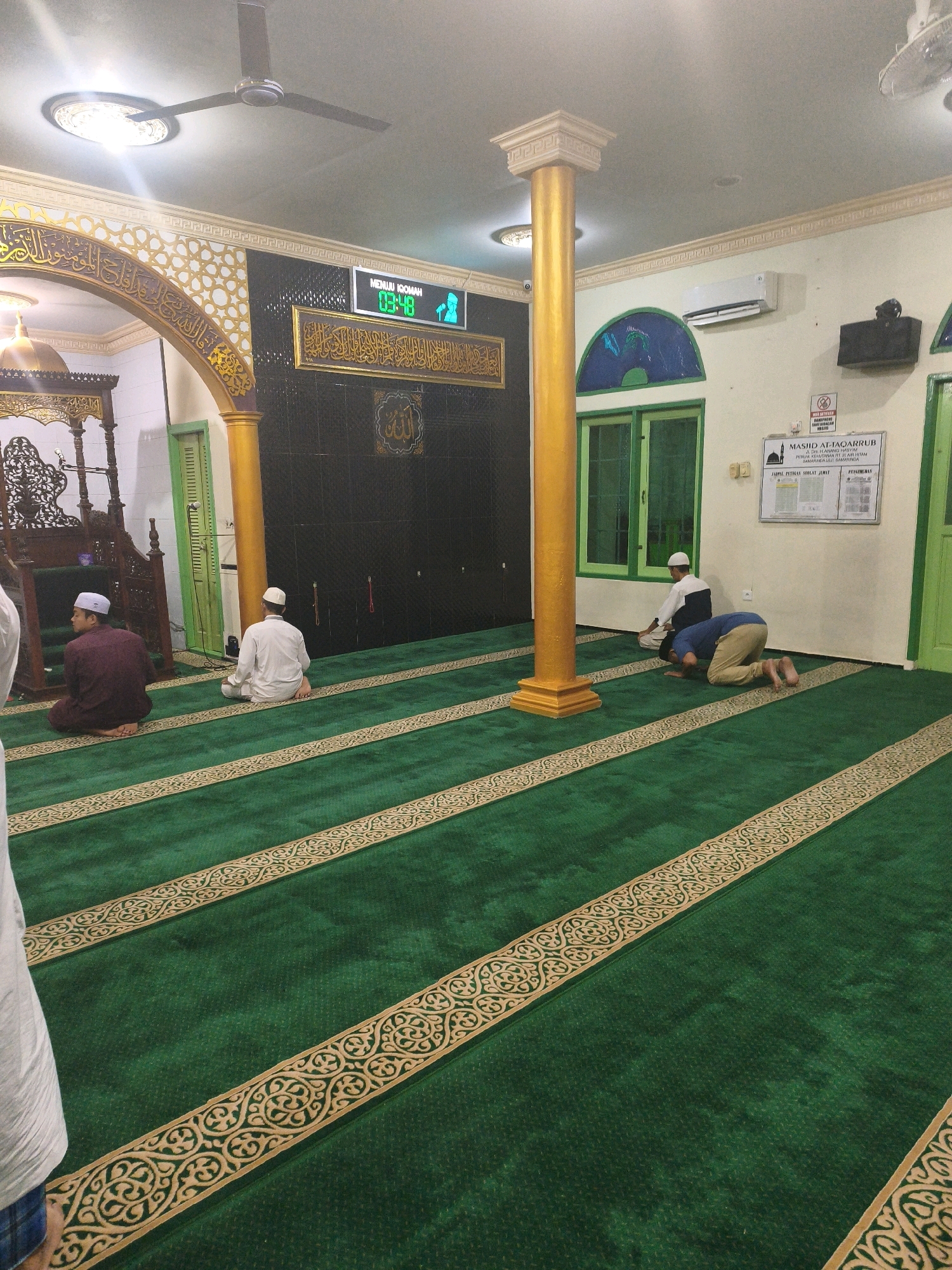 The image depicts the interior of the Masjid Attaqarrub in Samarinda, Indonesia. The mosque features a large, green carpet with ornate gold patterns demarcating prayer rows. In the foreground, a person wearing a white shirt and blue checkered pants is partially visible, standing on the carpet. Further into the mosque, three men are engaged in prayer. One man, dressed in a maroon robe and white cap, is seated on the carpet. Another man, in a white robe and cap, is also seated. A third man, wearing a blue shirt and tan pants, is in the prostration (sujud) position. The mosque's interior includes decorative arches, pillars painted yellow, a black wall adorned with Islamic calligraphy and a framed religious text, and a digital clock displaying "03:48" and "MENUI IQOMAH." There are green doors and windows visible, and an air conditioning unit mounted on the wall. A sign with the mosque's name and other information is also present. The lighting suggests it is either evening or a dimly lit interior.