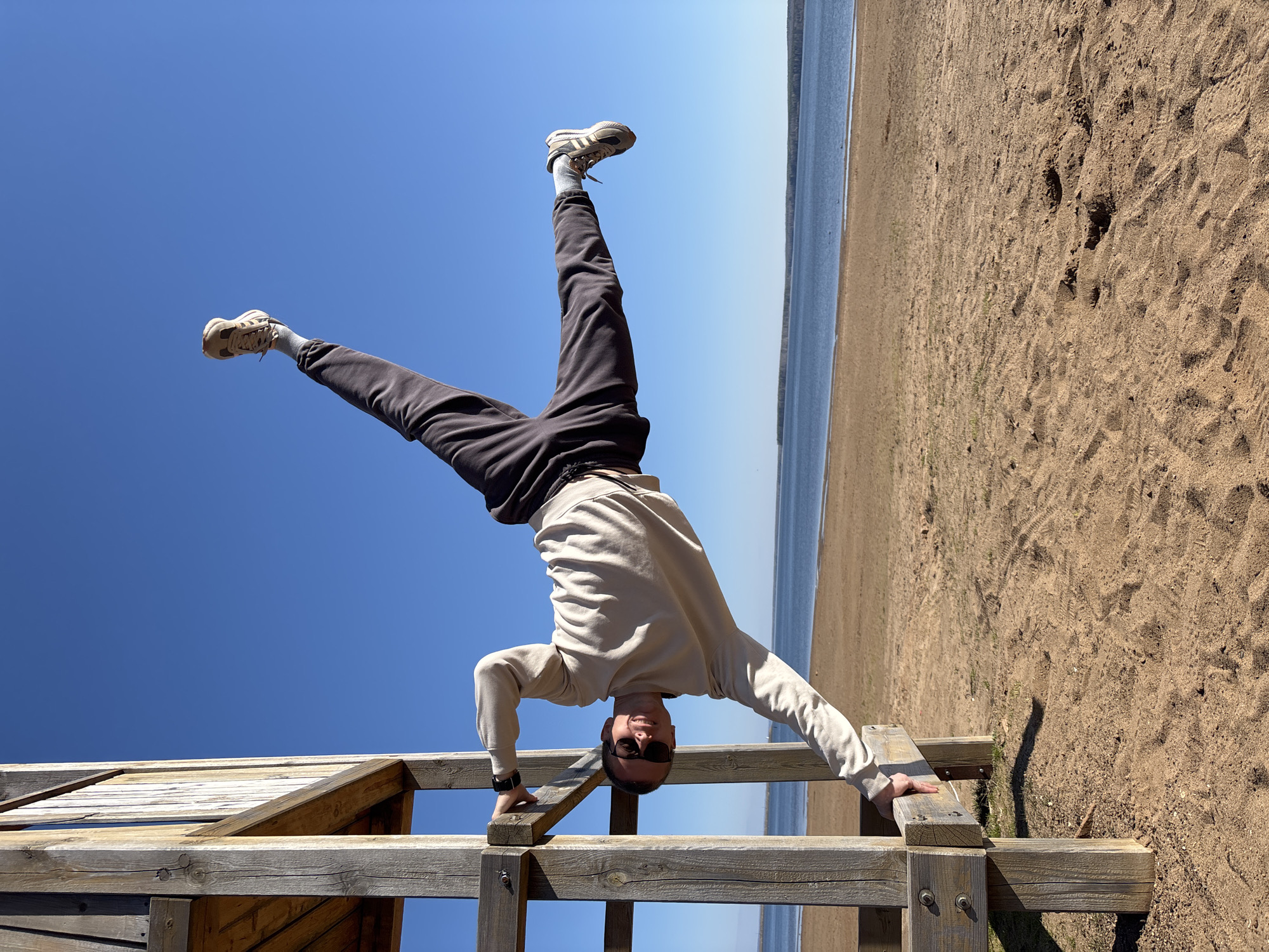A man is performing a handstand on a wooden structure. He is wearing sunglasses, a light-colored sweatshirt, dark pants, and sneakers. His legs are slightly spread apart and raised in the air. The background shows a clear blue sky, a sandy beach, and a body of water. The image is inverted, giving the impression that the man is upside down. The location is Chaikovsky, Russia, based on the metadata.