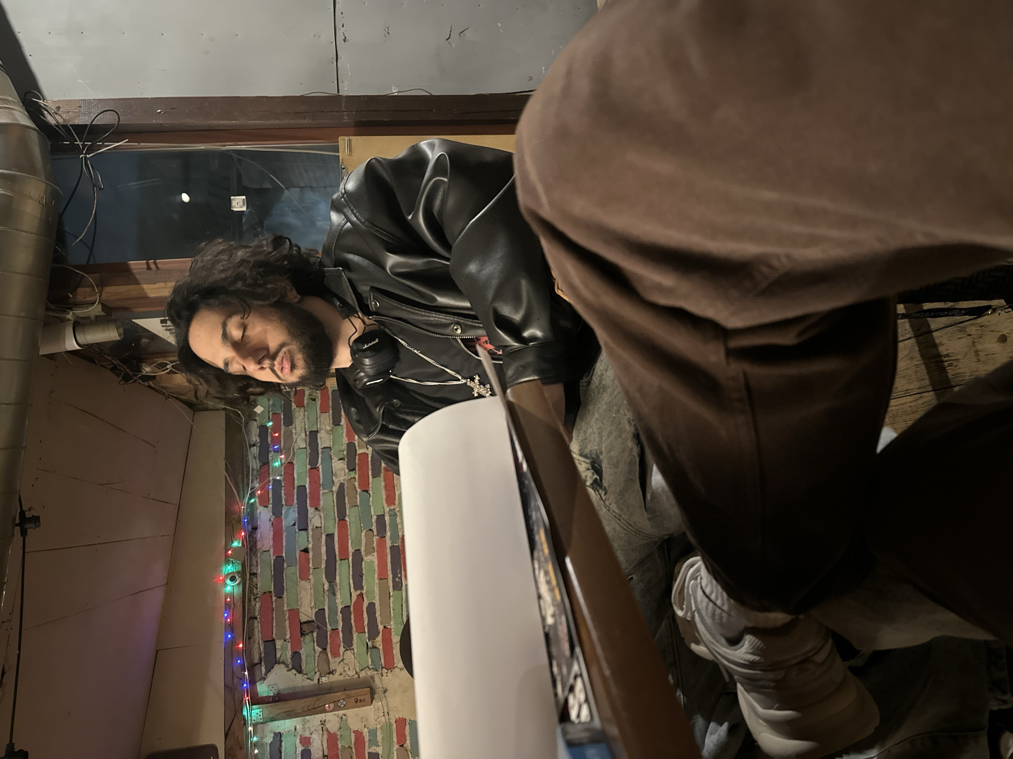 An interior scene depicts two individuals within what appears to be a casual establishment in Plovdiv, Bulgaria. In the mid-ground, a male subject with dark, curly hair and a beard is seated, his eyes closed and mouth slightly agape, suggesting repose. He is dressed in a black leather jacket, faded blue jeans, black over-ear headphones around his neck, and a silver chain with a prominent cross pendant. His left forearm rests on a wooden surface where a large white roll of material and printed items are present. Partially visible in the immediate foreground are the lower legs of a second seated individual, clad in brown pants and light-colored sneakers. The background features a multi-colored brick wall (red, blue, green, purple, beige) adorned with a string of multi-colored LED fairy lights. Overhead, exposed metal ductwork and electrical wiring are visible against a light-toned plank ceiling. To the right, a dark exterior is visible through a window or opening, marked by faint distant light and utility wiring. The overall lighting is dim.
